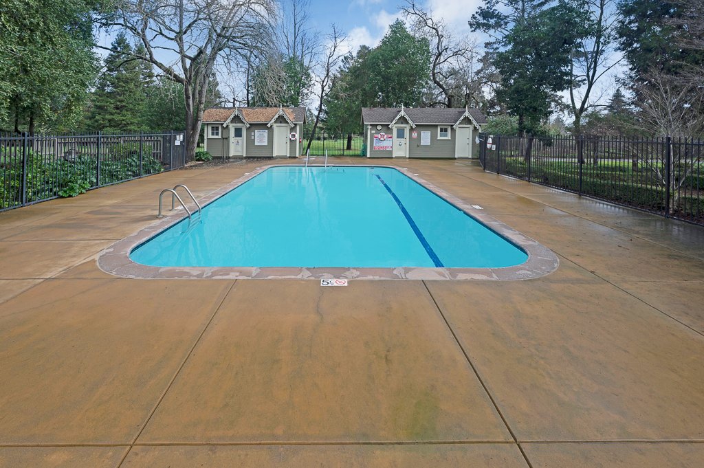 Empty outdoor swimming pool with a blue liner is surrounded by a wet concrete deck, fenced by black metal railings, with small buildings and trees in the background under a partly cloudy sky.