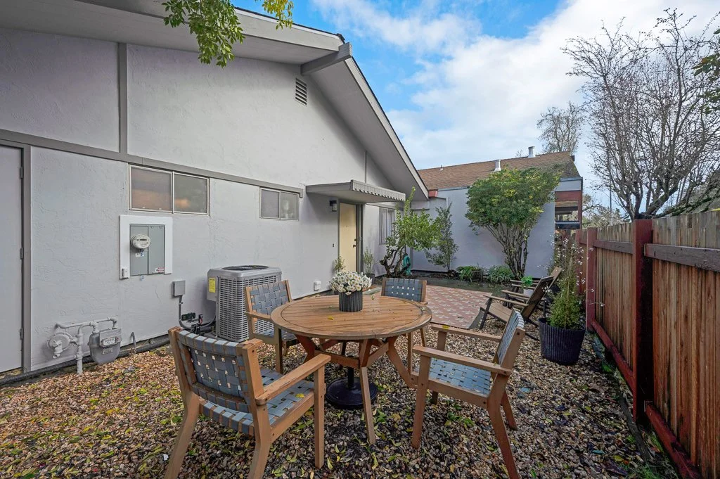 Backyard patio with a round wooden table and four chairs, surrounded by trees, plants, and a wooden fence, with a white house in the background.