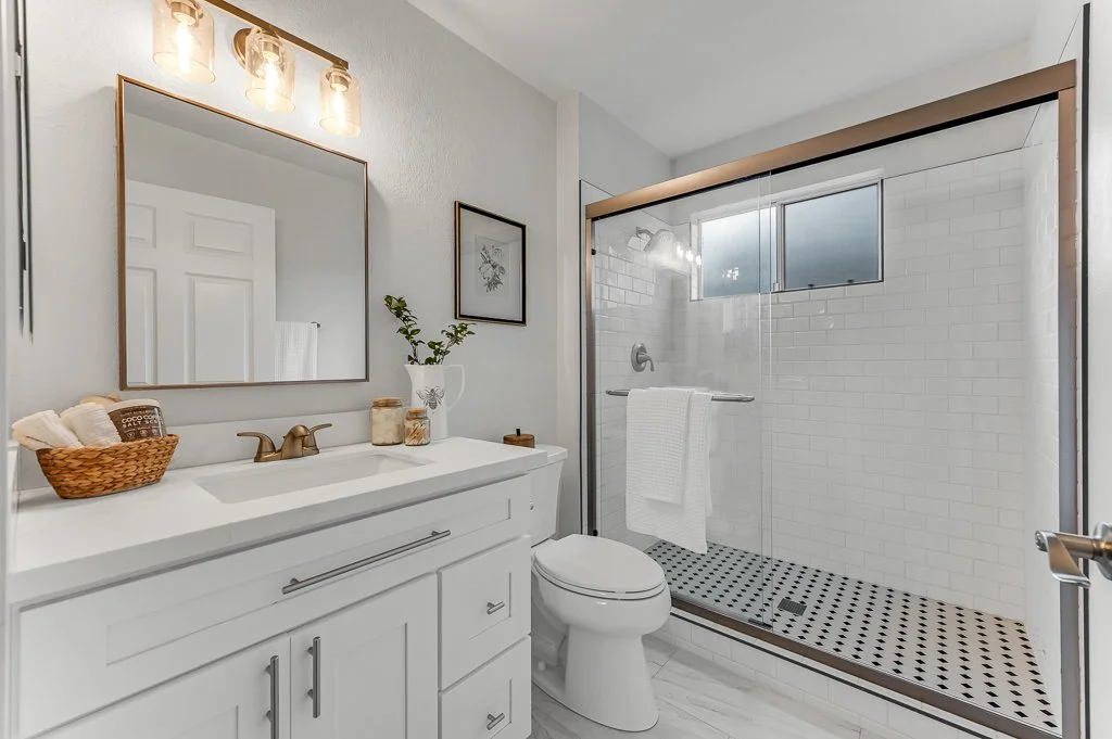Modern bathroom with white vanity, large mirror, vanity lights, potted plant, framed art, toilet, and walk-in shower with glass door and tiled wall.