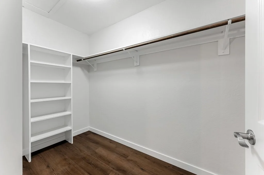 Empty walk-in closet with white walls, wooden flooring, a white shelving unit on the left, and a wooden clothing rod supported by white brackets on the right wall.