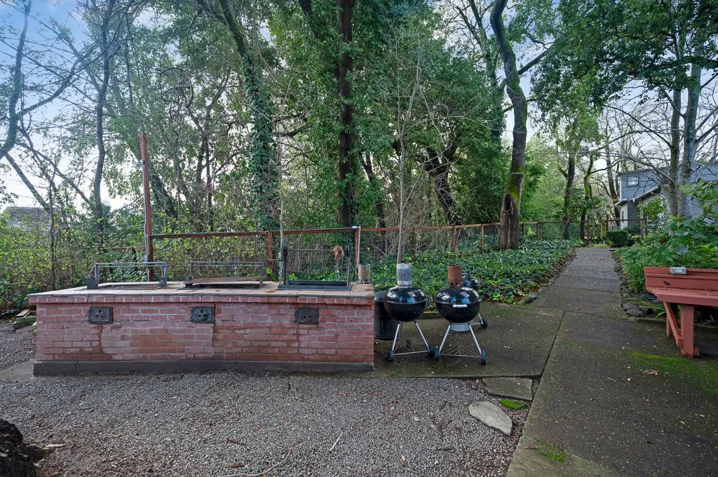 Outdoor backyard with brick grilling station, two small black charcoal grills, and a paved walkway leading through trees and greenery.