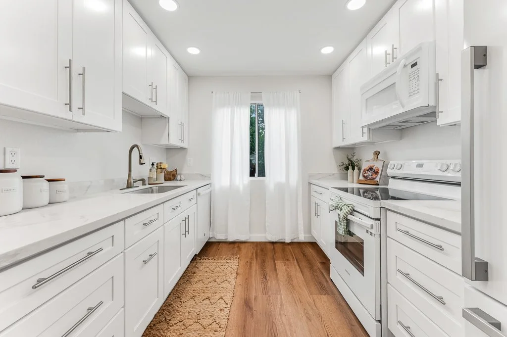 Bright white kitchen with white cabinets, white appliances, a window with white curtains, and wooden flooring.