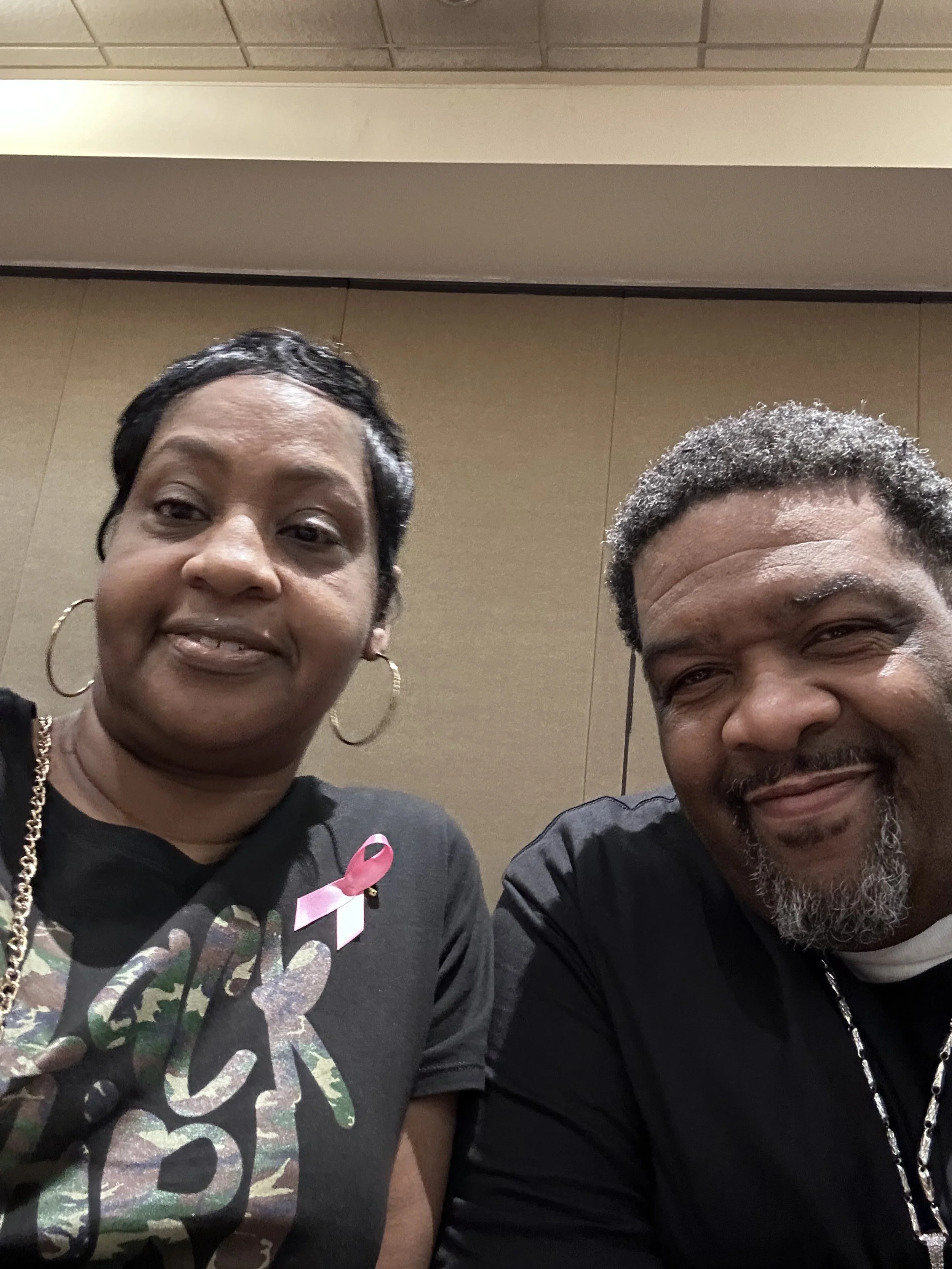 A woman and a man taking a selfie together indoors. The woman is wearing a black shirt with pink ribbon and camouflage parts, has short black hair, and is wearing hoop earrings. The man has short curly gray hair, a beard, and is also wearing a black shirt.