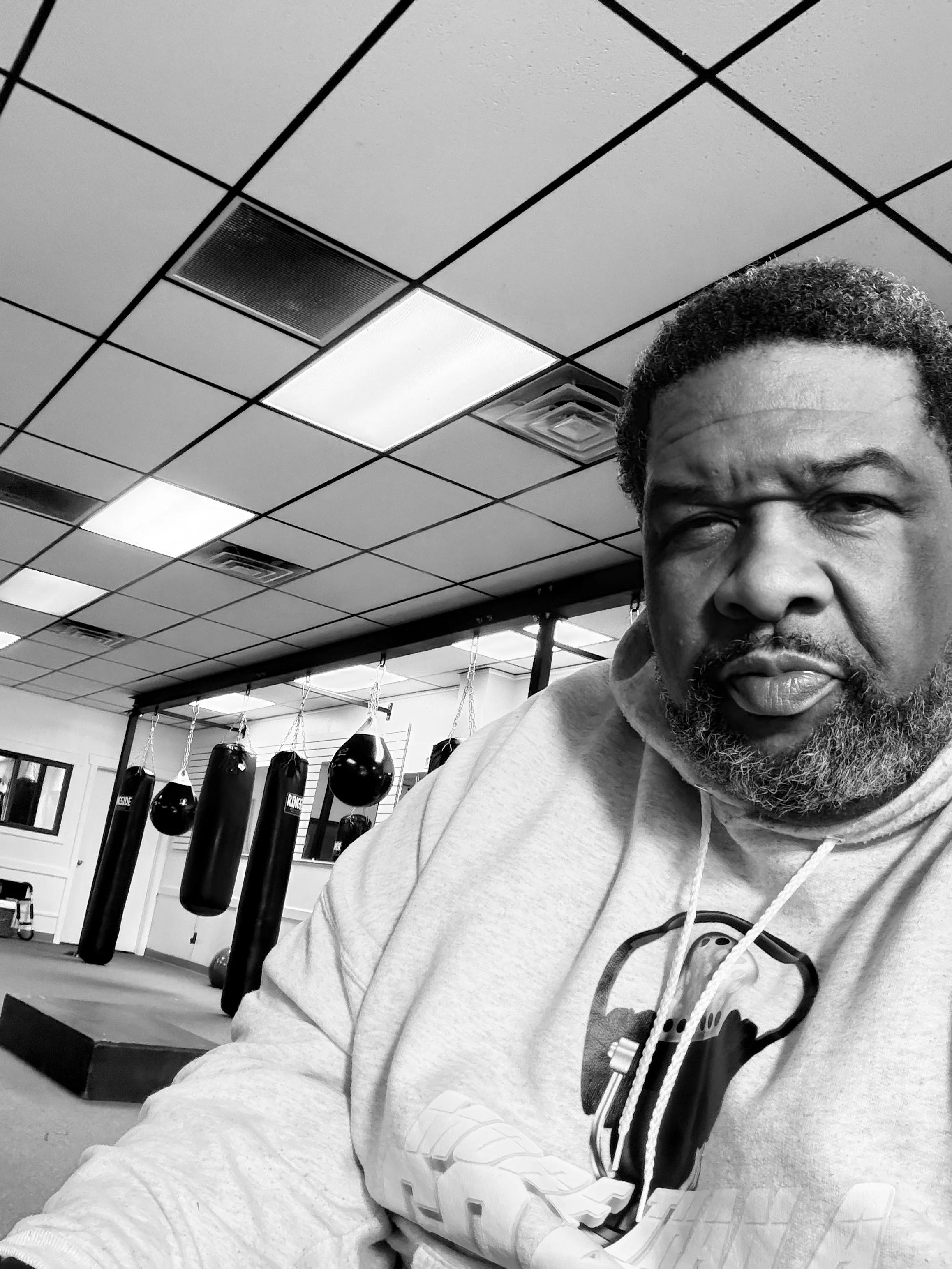 A man with a beard and curly hair taking a selfie in a gym with boxing punching bags in the background.