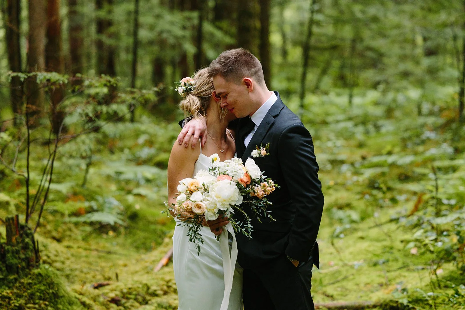 A bride and groom embracing in a forest, with the bride holding a bouquet of flowers. Florist for weddings in Stroud, Wotton Under Edge, Bristol, Yate, Dursley, Tetbury, Malmesbury and Forest of Dean. 