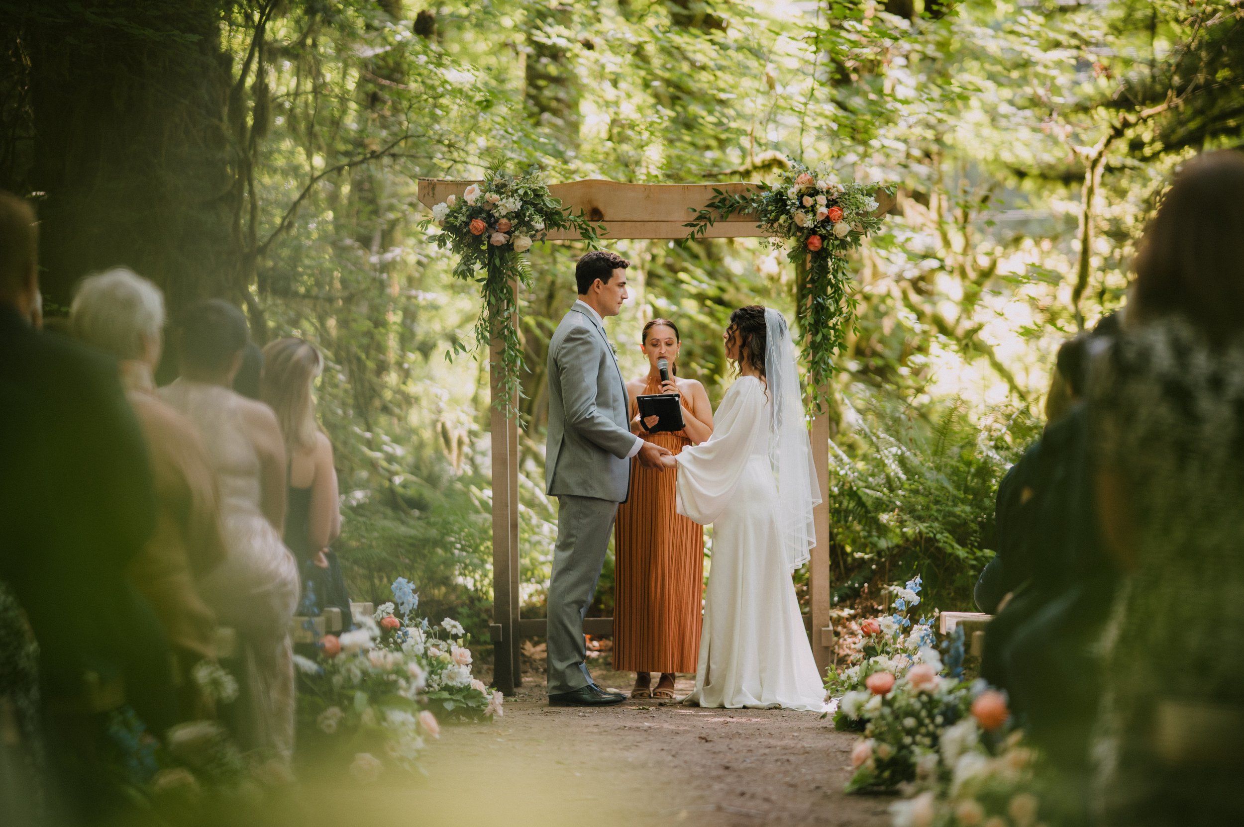 A wedding ceremony taking place outdoors in a lush green forest, with a bride and groom holding hands and facing each other under a wooden arch decorated with flowers. An officiant stands between them, aisle flowers are either side 