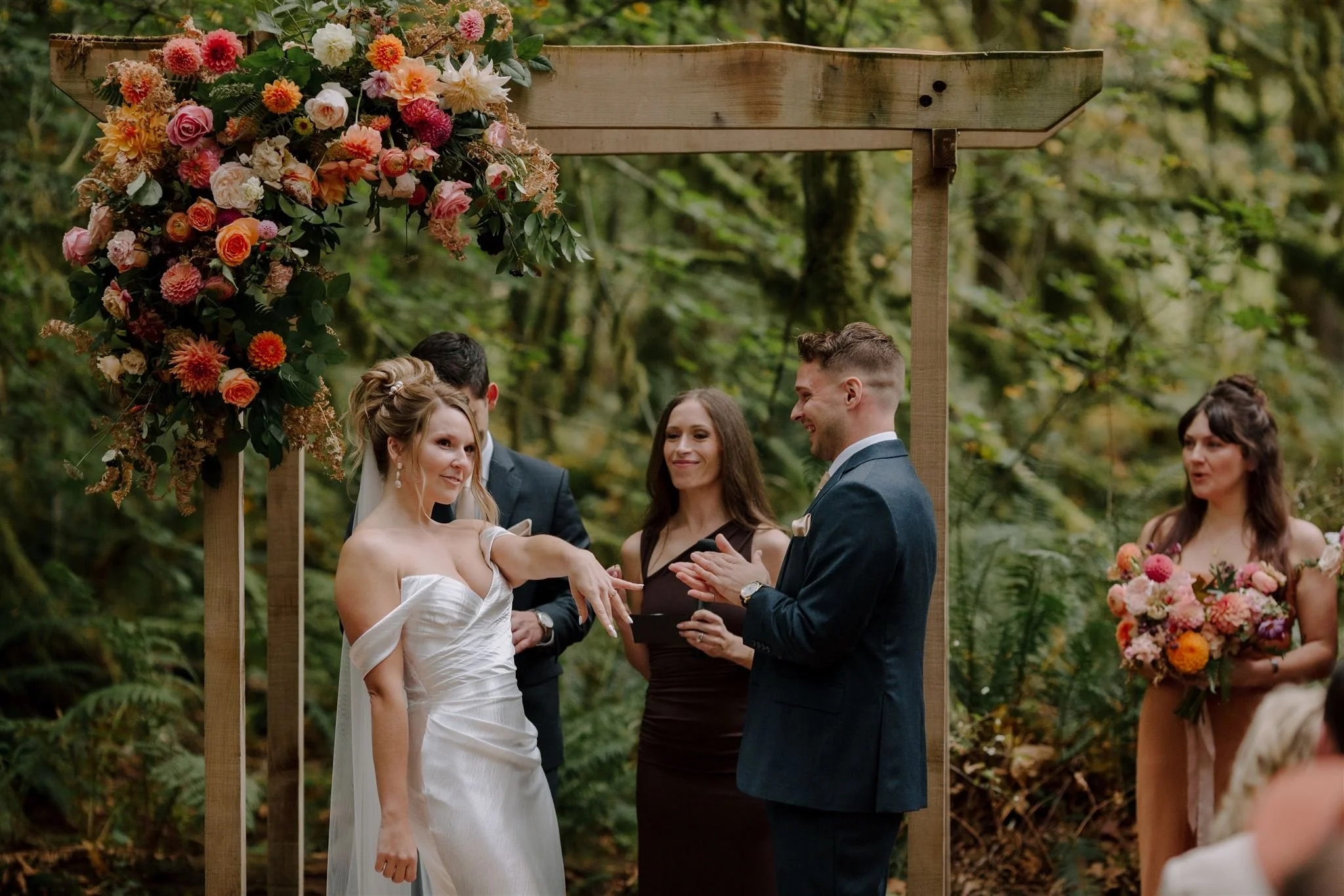 Wedding ceremony with a bride and groom exchanging vows under a wooden arch decorated with flowers, in a lush forest setting.