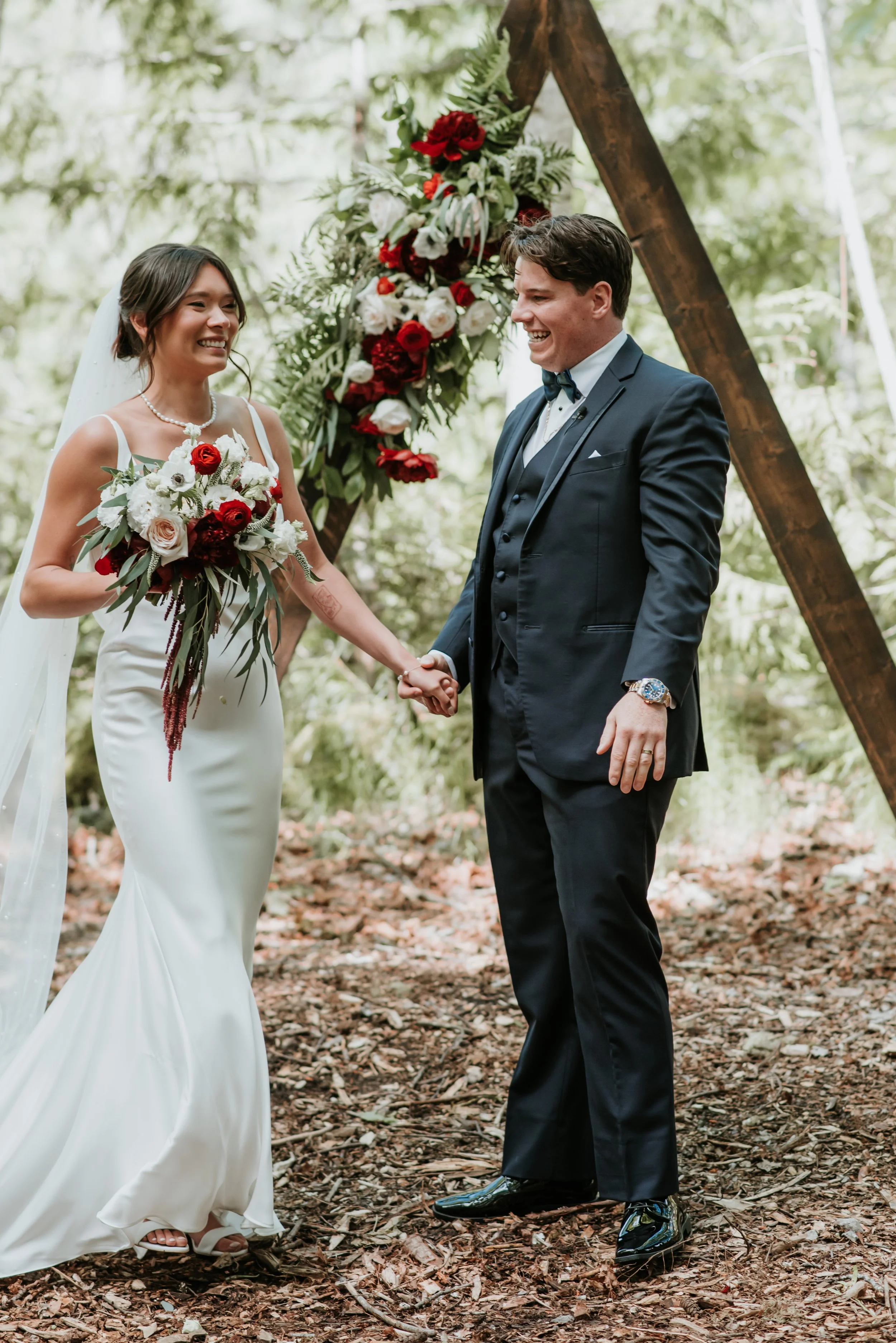 A bride and groom holding hands during a wedding ceremony outdoors in a wooded area, standing in front of a wooden arch decorated with red and white flowers and greenery, smiling at each other.