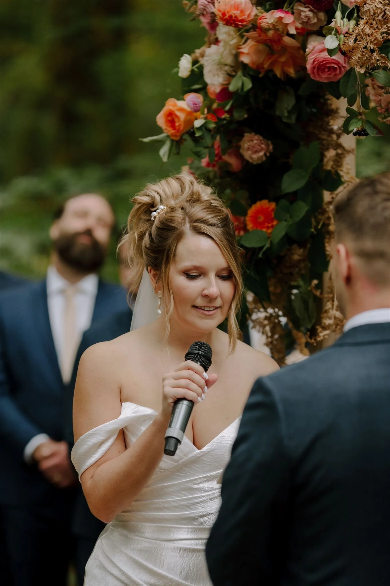 A woman in a white off-shoulder wedding dress holding a microphone, exchanging vows during an outdoor wedding ceremony. A floral arch with pink, orange, and white flowers is in the background.Florist for weddings in Stroud, Wotton Under Edge, Bristol