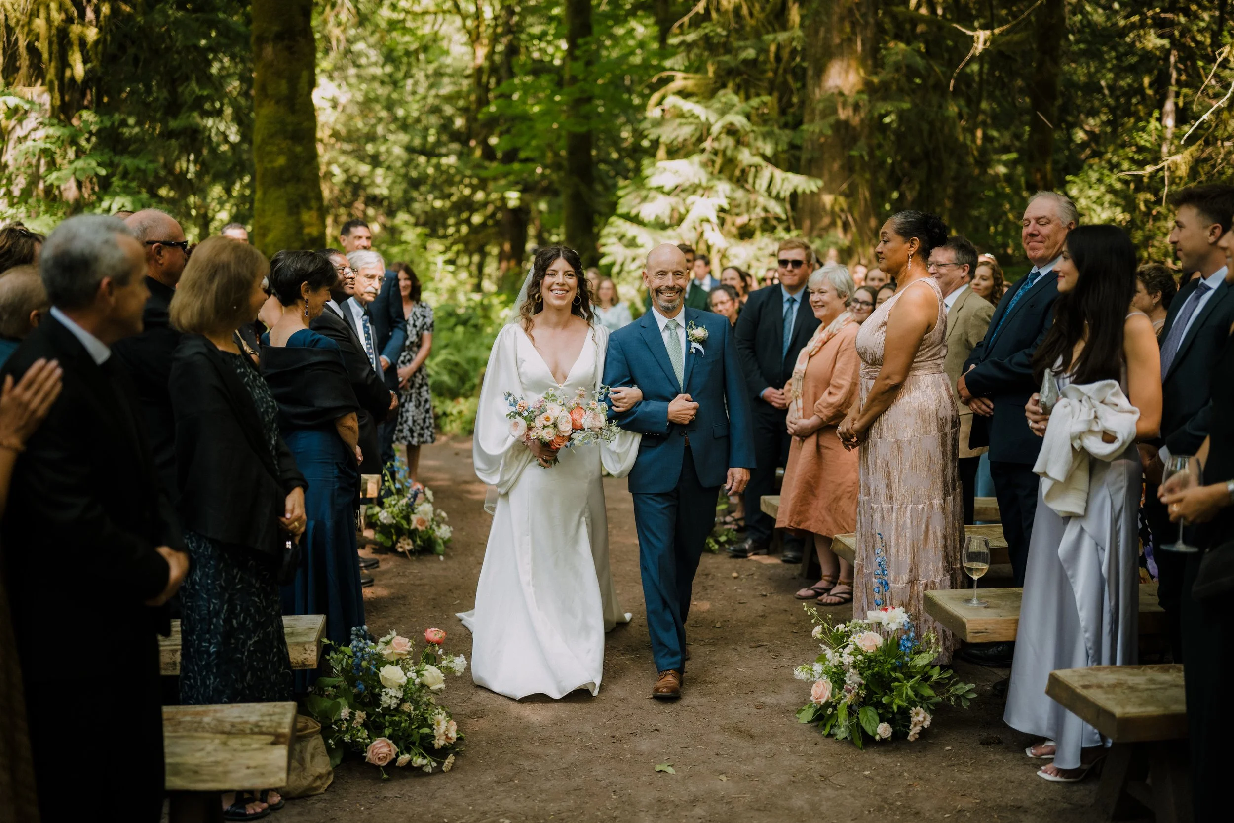 A wedding ceremony in a forest with a bride in a white dress holding a bouquet walking down the aisle with a man in a blue suit. Guests are standing on either side, smiling and watching, surrounded by greenery and flowers.