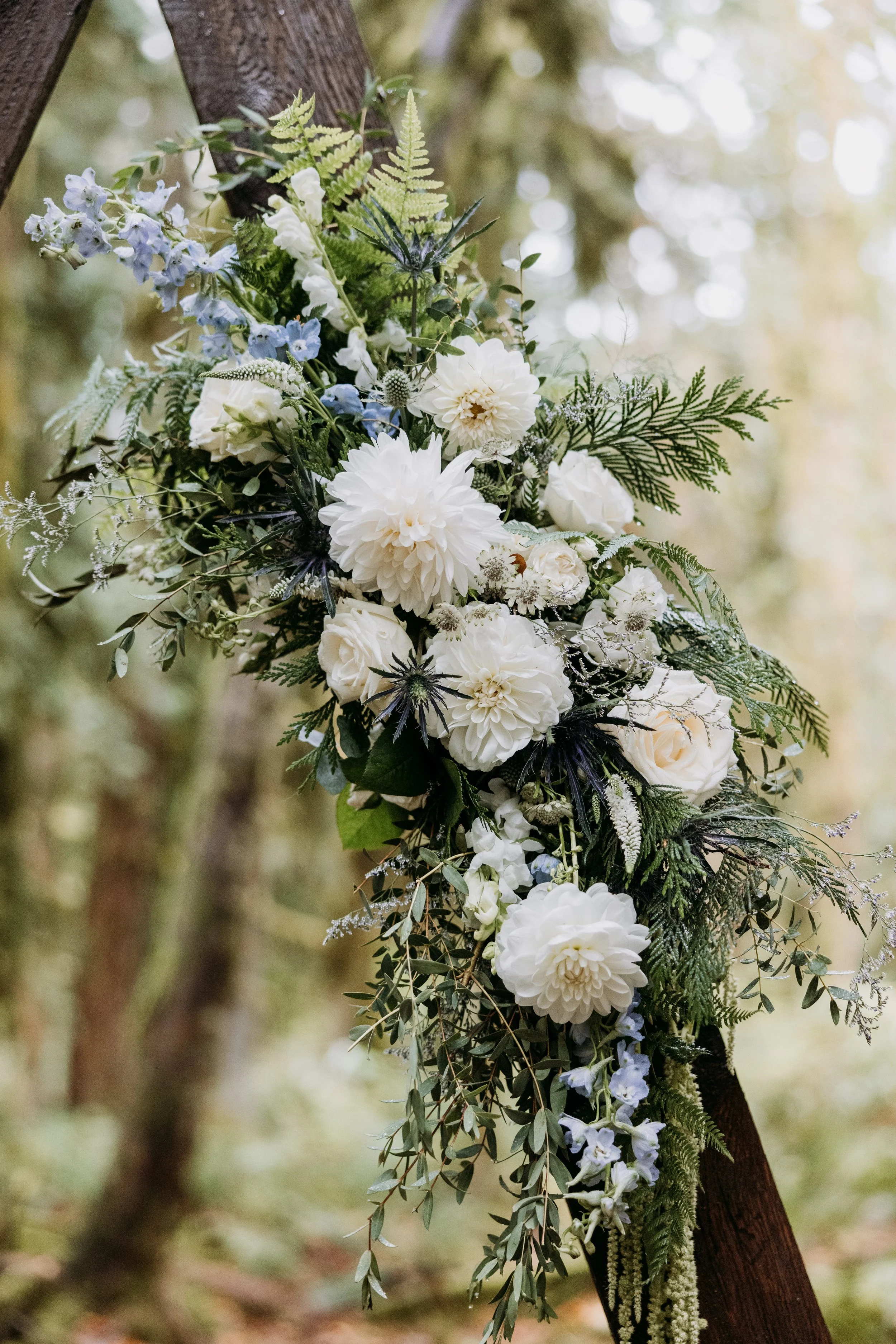 A floral arrangement with white dahlias, roses, and other flowers, attached to a wooden structure outdoors with a blurred forest background.