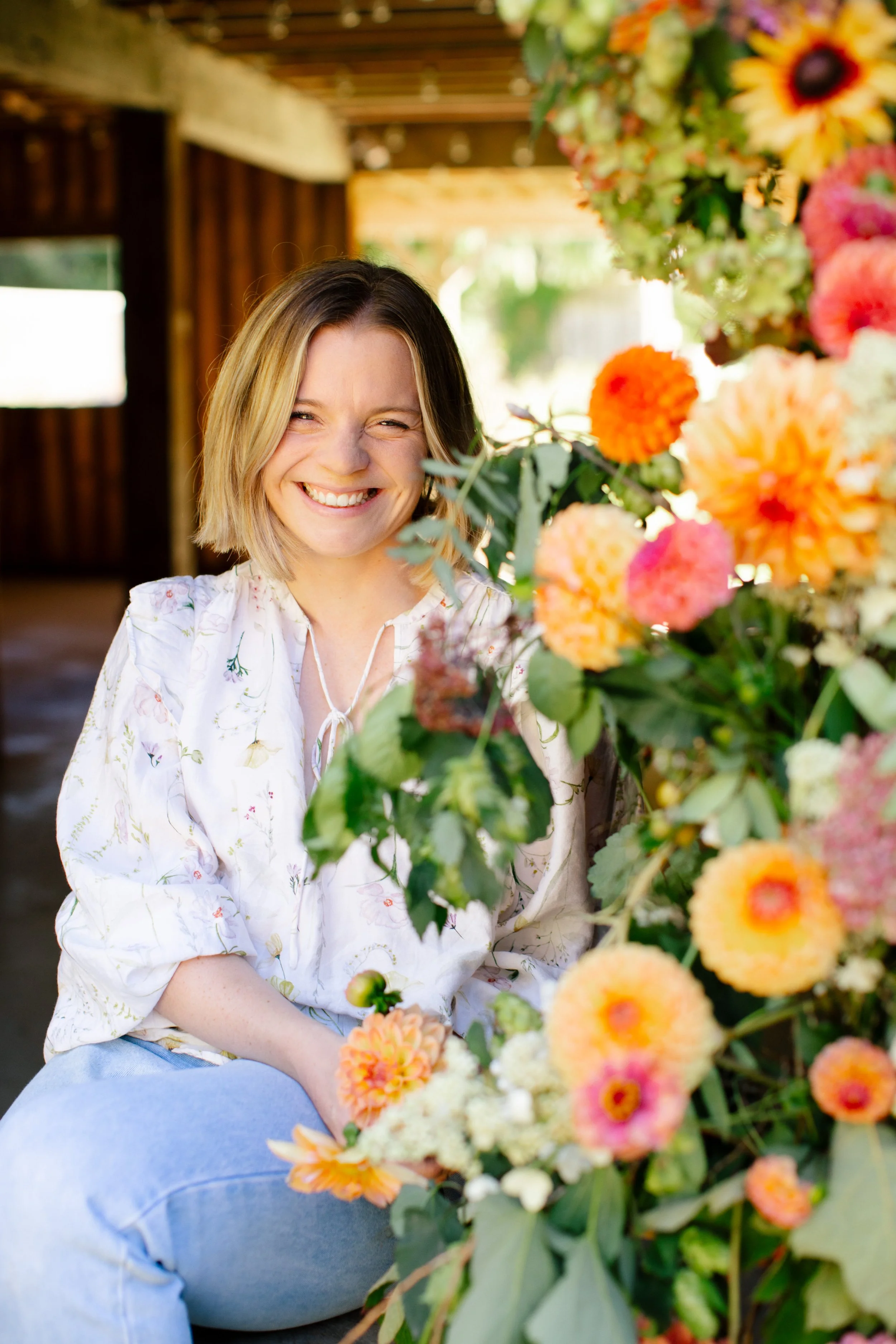 A woman with shoulder-length brunette and blonde hair smiling with eyes closed, wearing a white floral blouse and blue jeans, sitting amidst colorful flowers with orange, pink, white, and green foliage, in a rustic outdoor setting with wooden beams