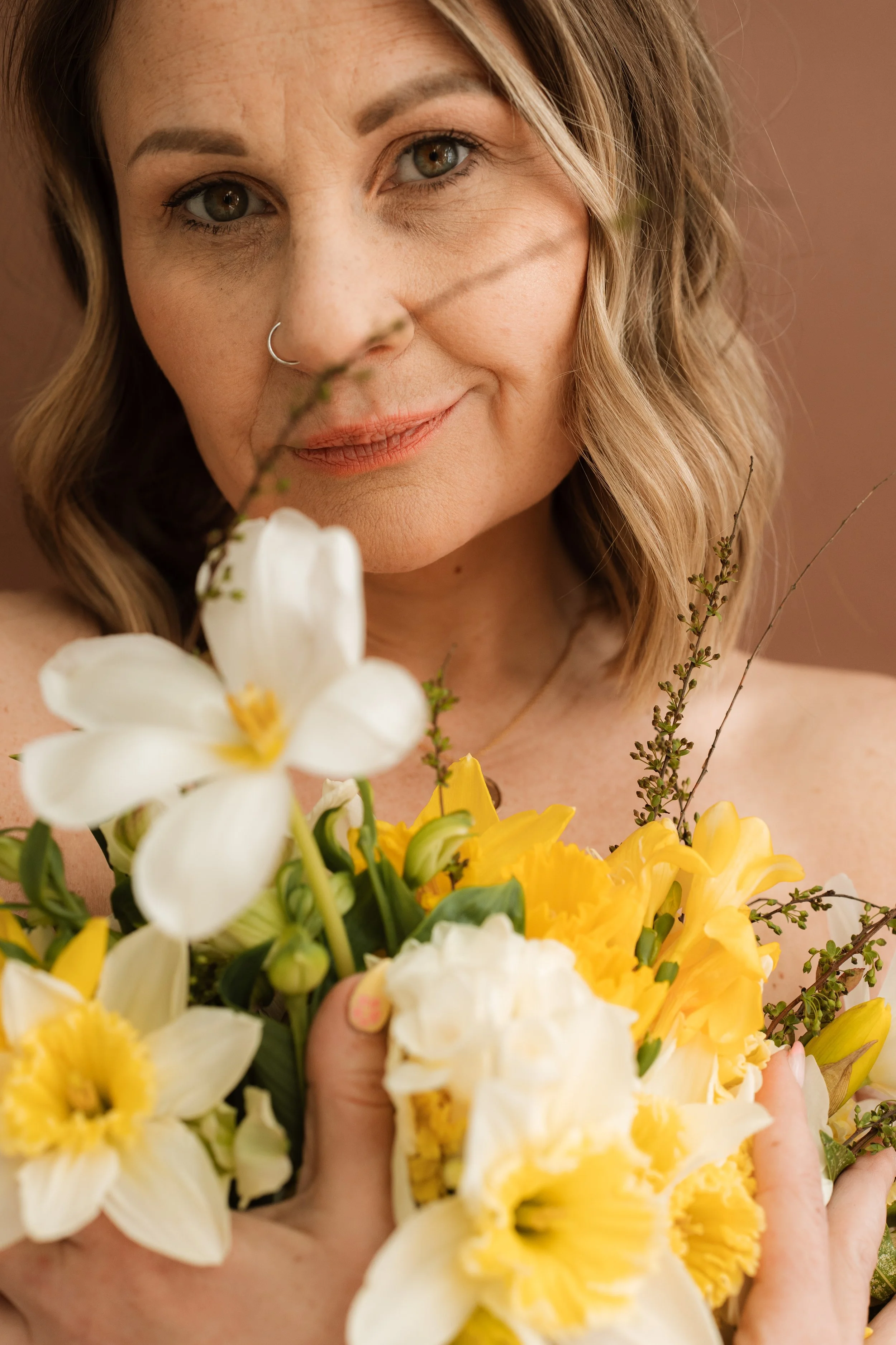 A woman with wavy blonde hair holds a bouquet of yellow and white flowers close to her chest, smiling softly at the camera.