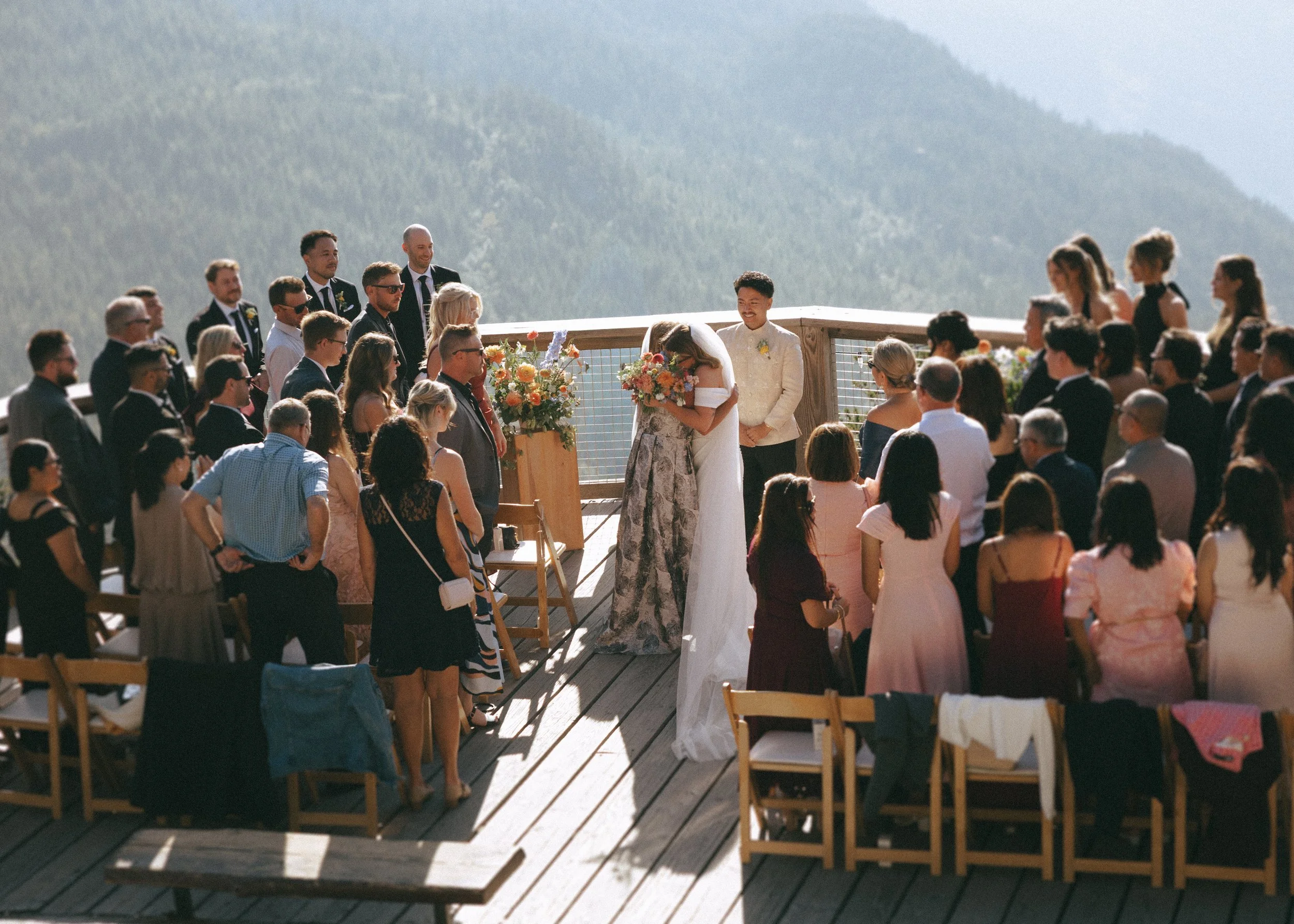 A group of people gathered for a wedding ceremony outdoors on a wooden deck with a scenic mountain backdrop. The bride and groom are standing together, surrounded by friends and family.