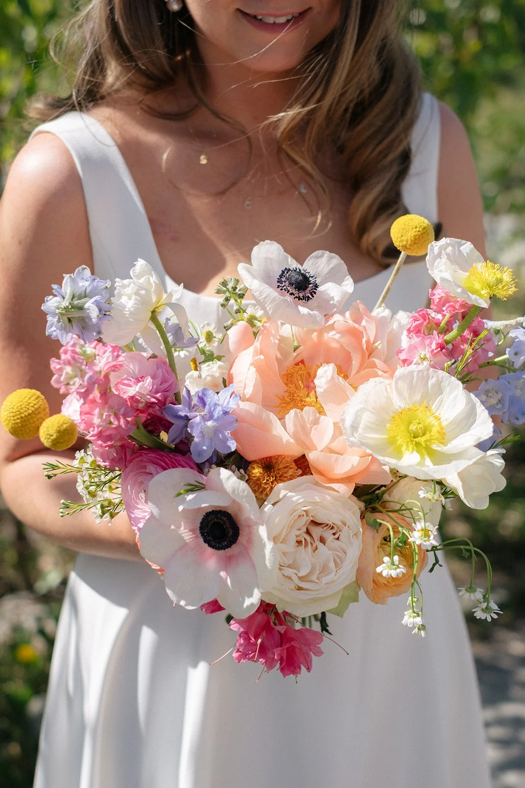 A woman in a white dress holds a vibrant bouquet of various colorful flowers, including white, pink, peach, yellow, and purple blossoms, in an outdoor setting.