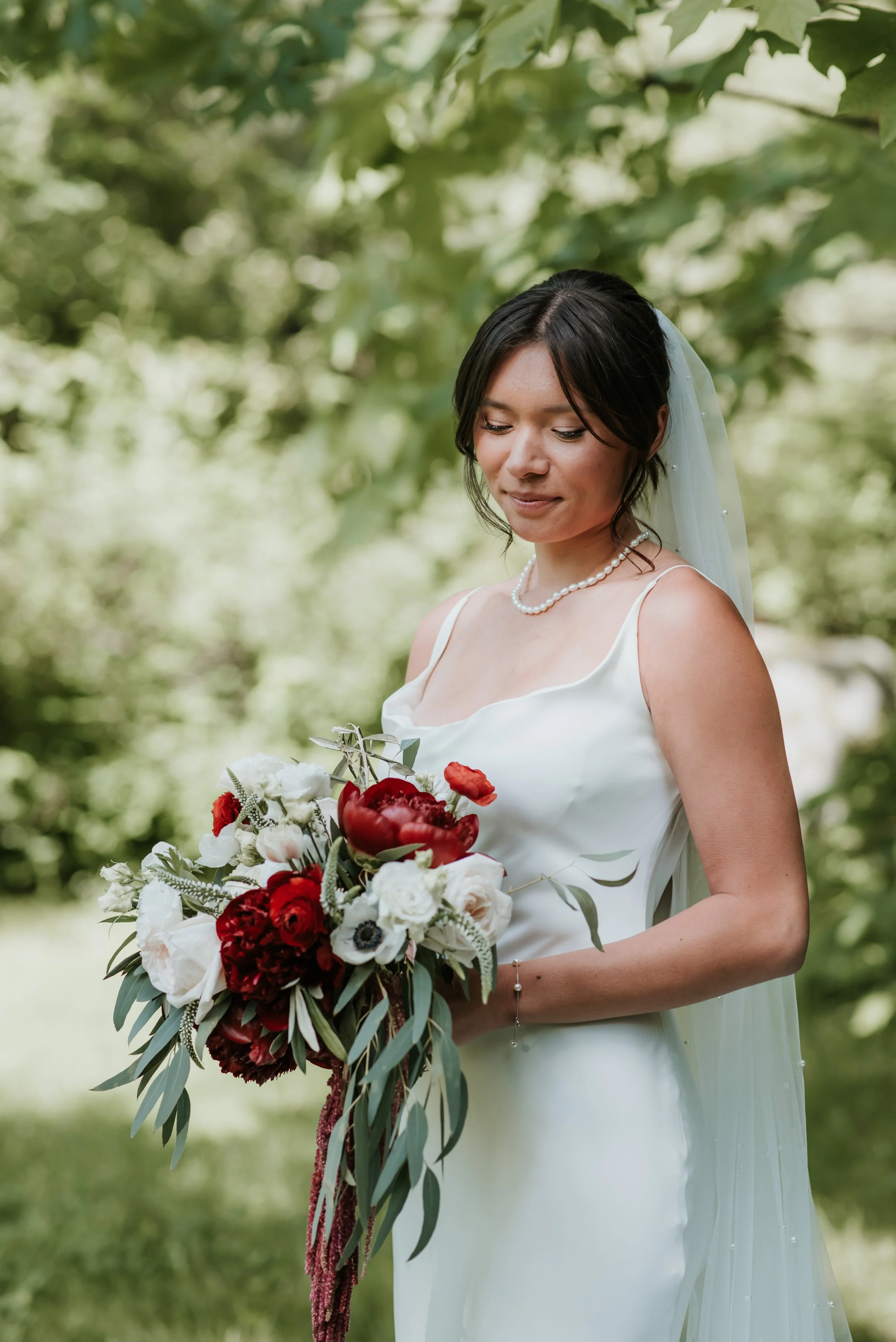 A bride outdoors holding a bouquet of red, white, and pink flowers with greenery, wearing a white wedding dress, pearl necklace, and veil, with green leaves in the background. Florist for weddings in Gloucestershire
