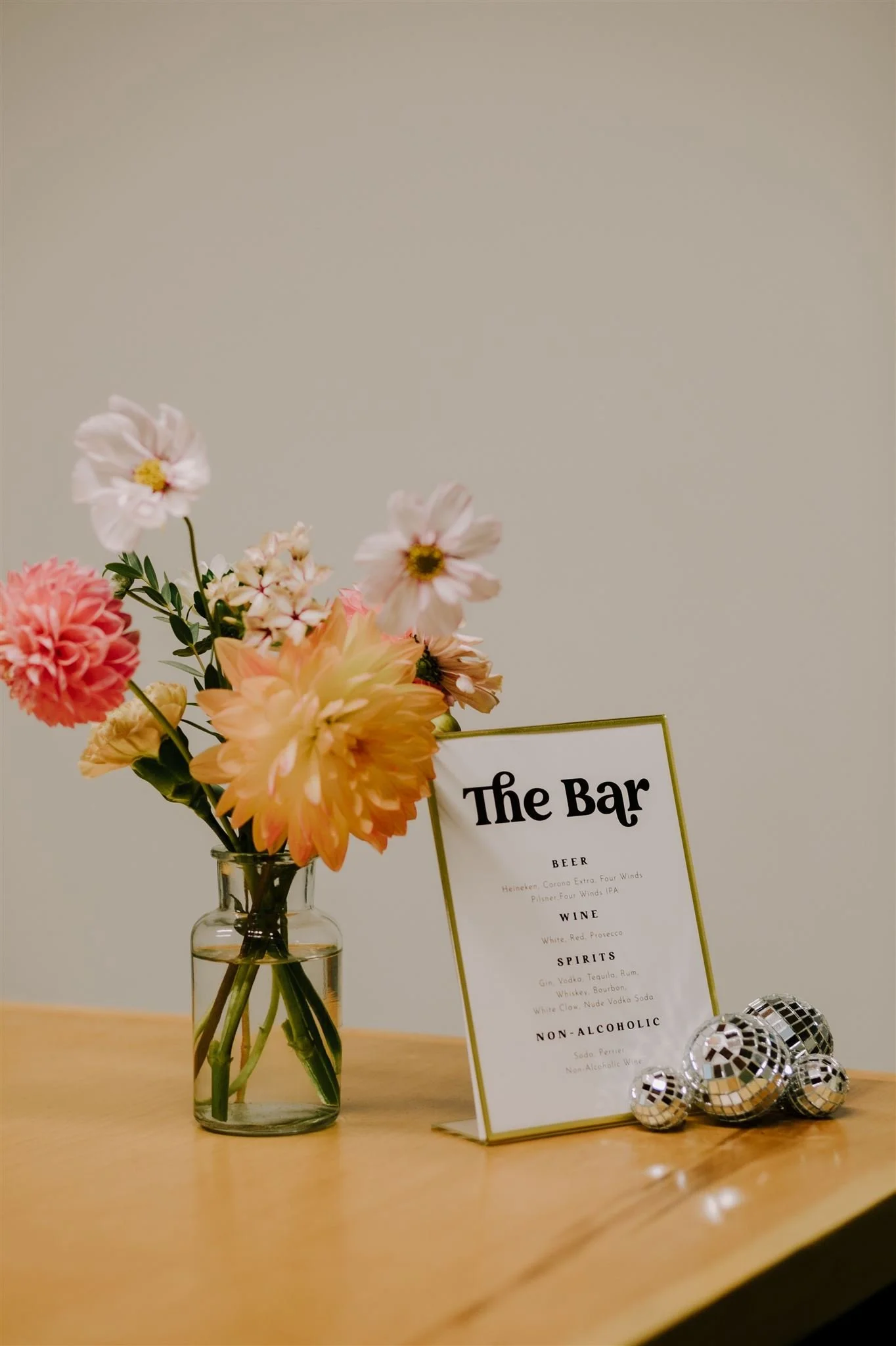 A bud vase of seasonal flowers in a glass vase on a wooden table, next to a menu for a bar featuring drink options including beer, wine, spirits, and non-alcoholic beverages, and decorated with small disco balls.