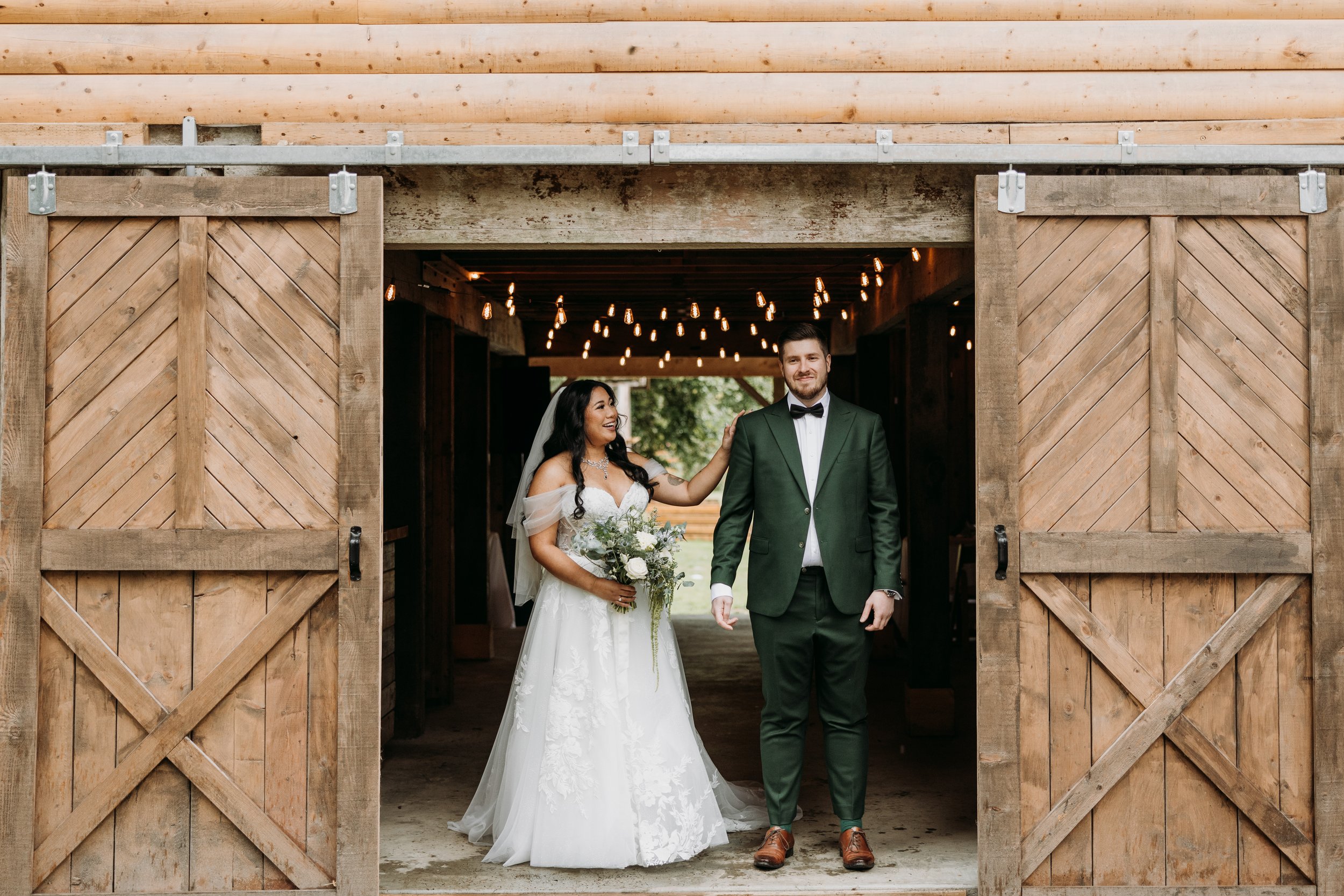 A bride and groom inside a rustic wooden barn, smiling, with string lights hanging overhead. The bride is holding a bouquet of white flowers, and the groom is wearing a dark green suit with a bow tie. Florist for weddings in Stroud, Wotton Under Edge