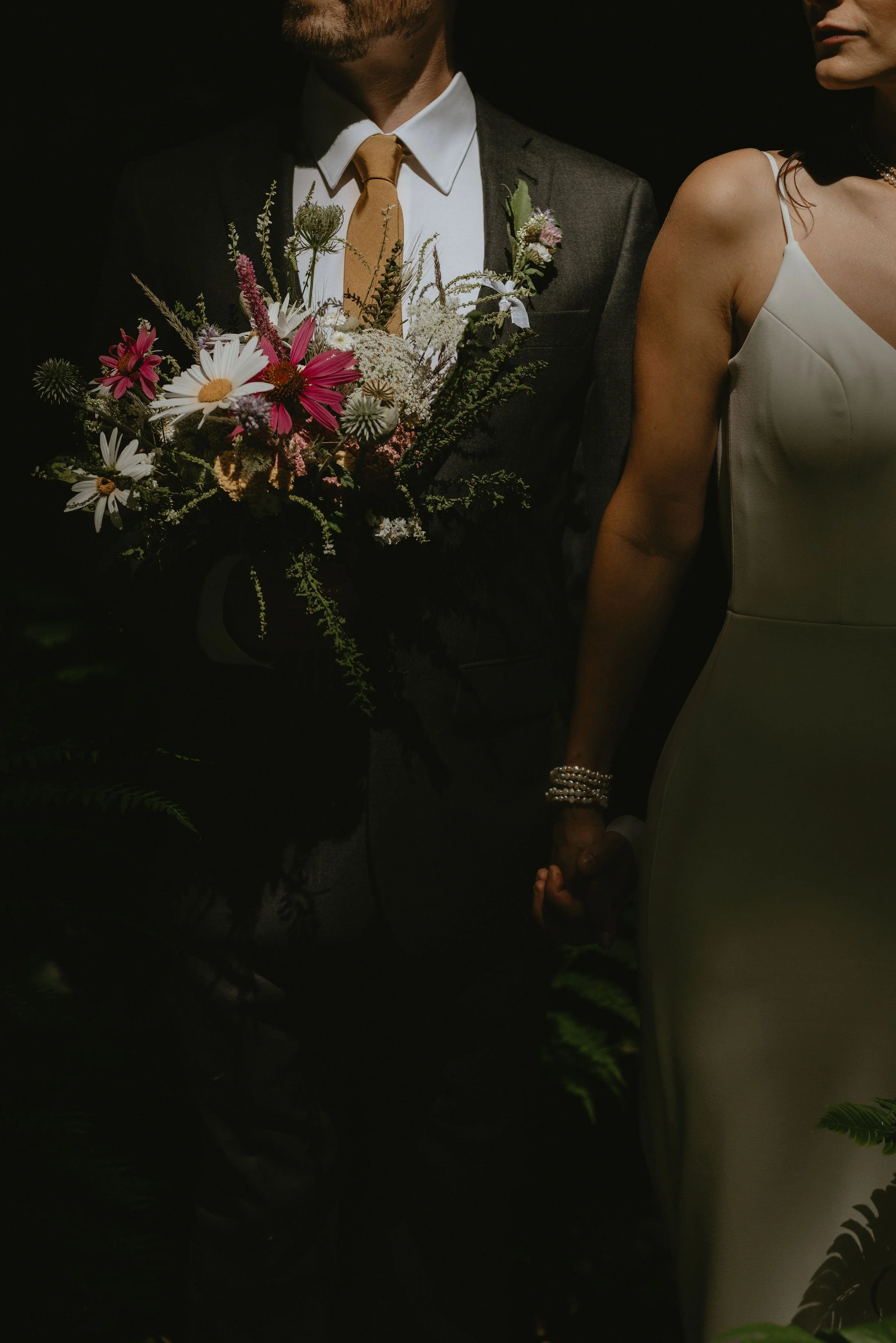 A close-up photo of a newlywed couple holding hands, with the groom in a suit and tie holding a bouquet of colorful flowers. The bride is wearing a white dress and jewelry, and they are partially obscured by shadows and dark surroundings.