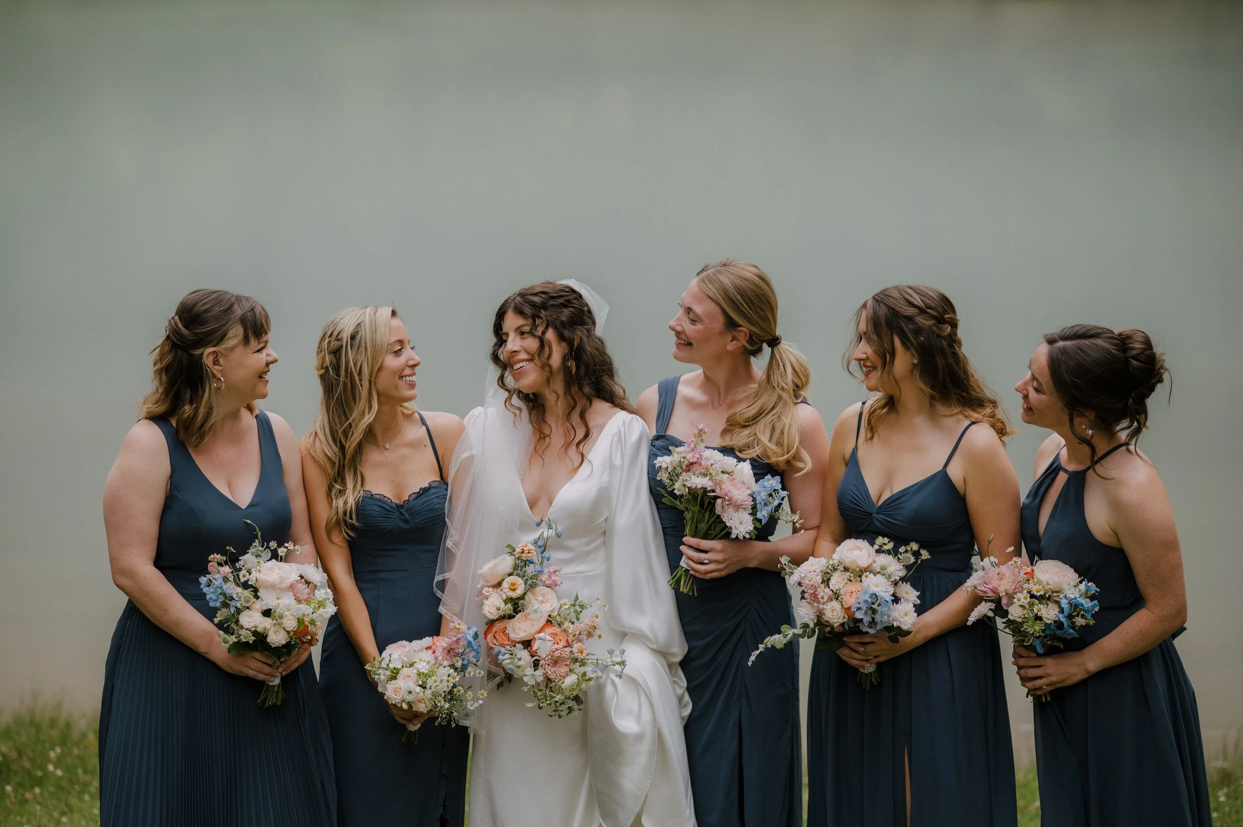 A bride in white holding a bouquet surrounded by six bridesmaids in dark blue dresses, all holding floral bouquets, standing outdoors against a plain green background.