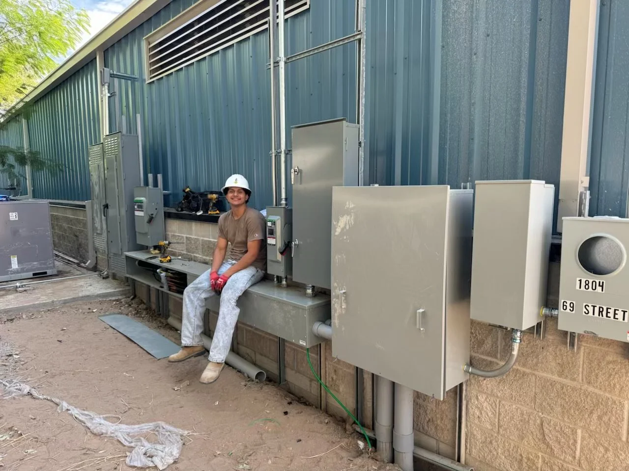 A woman sitting on a metal utility box outside a building with electrical and mechanical equipment, wearing a white hard hat, work gloves, and work clothes.
