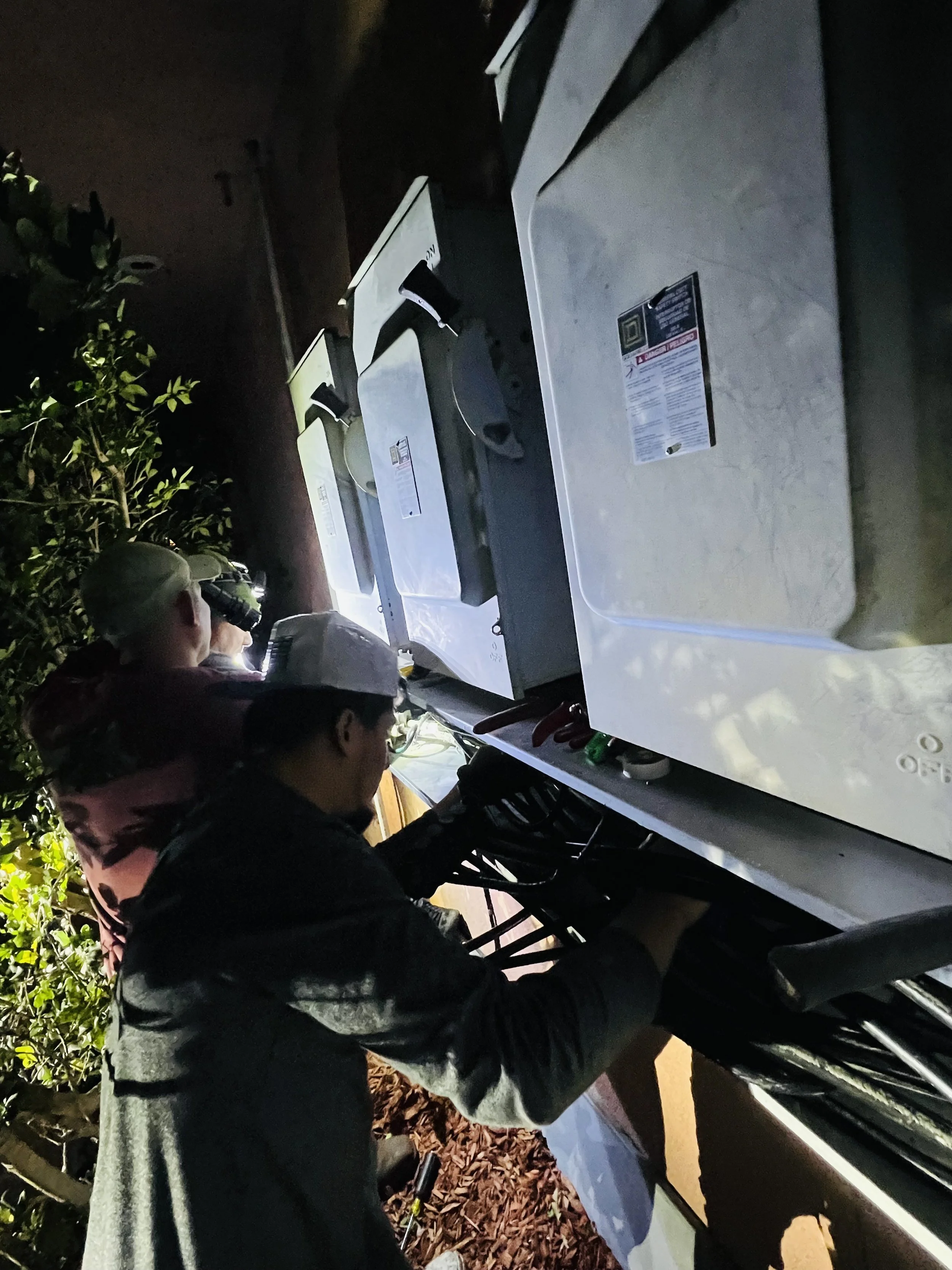 People working with utility meters or electrical equipment outdoors at night.