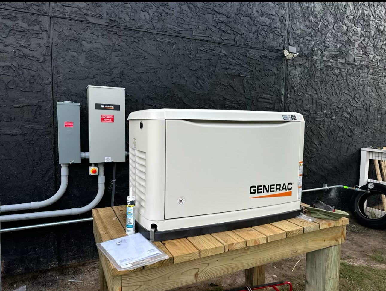 A Generac standby generator installed on a wooden platform outside, near a black textured wall, with electrical panels and wiring nearby.
