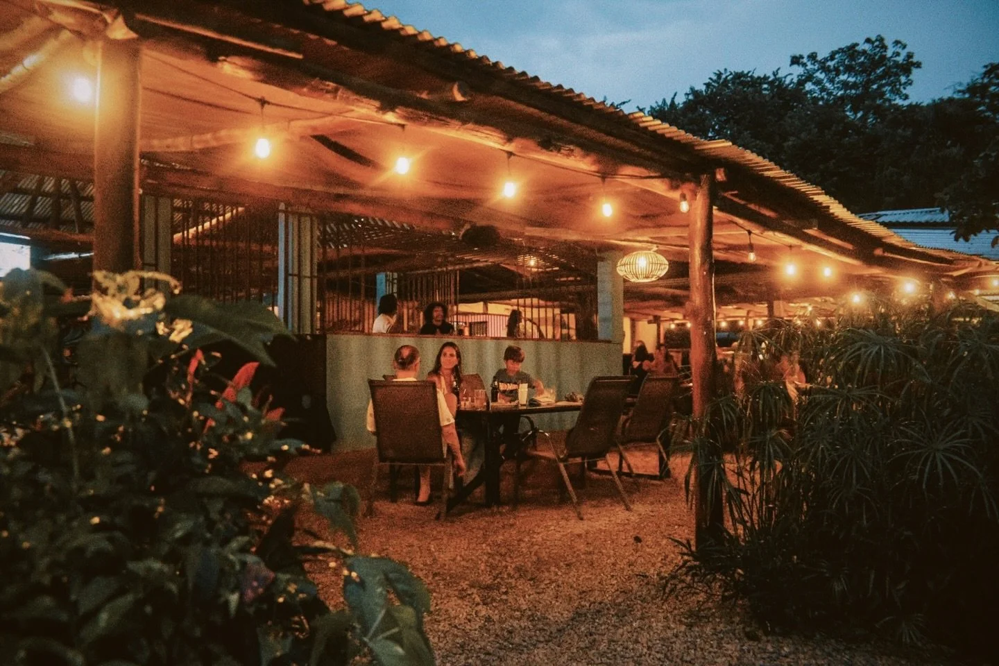 An outdoor dining area illuminated with warm lights at dusk or evening. Several people are seated at tables, enjoying their meals, while the background features a rustic wooden structure with string lights and greenery.