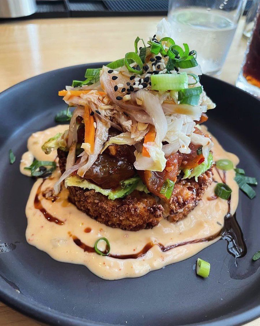 A fried dish topped with shredded vegetables, sliced green onions, black and white sesame seeds, and a drizzle of sauce, served on a bed of creamy sauce on a black plate.