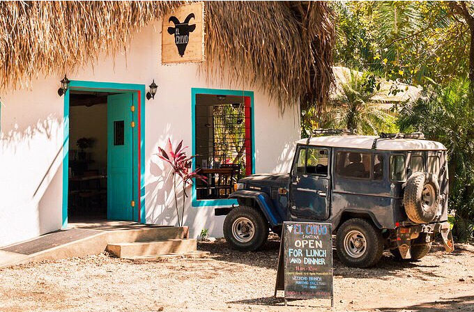 A small white building with a thatched roof, turquoise trim around the door and window, and a sign reading 'El Chivo' above the door. A vintage black off-road vehicle parked in front, and an A-frame chalkboard sign advertising the place is open for lunch and dinner.