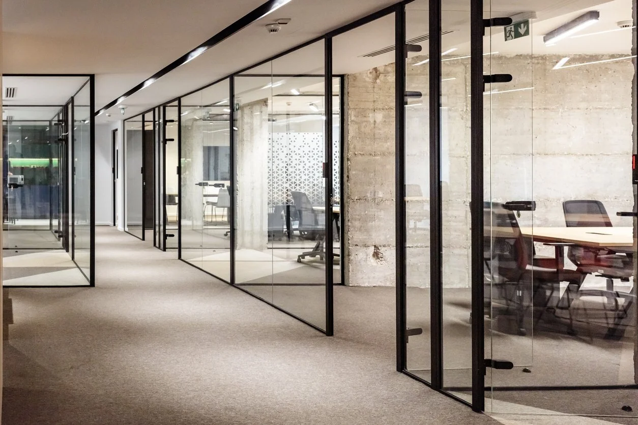 Modern office corridor with glass-walled meeting rooms and workspaces, beige carpet, and minimalistic design.