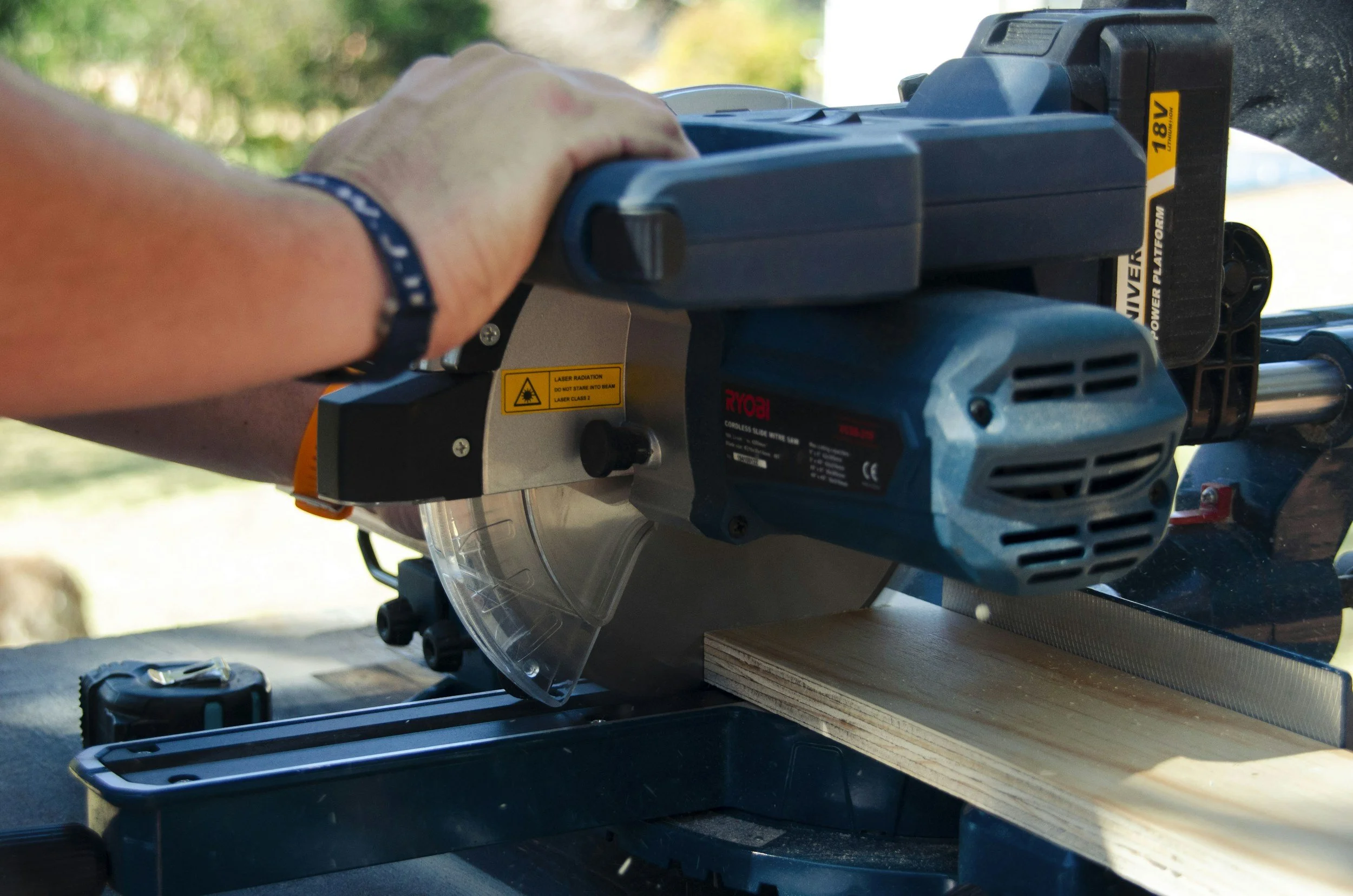 Close-up of a person's hand operating a Ryobi cordless miter saw on a wooden piece, set outdoors with a blurred green background.
