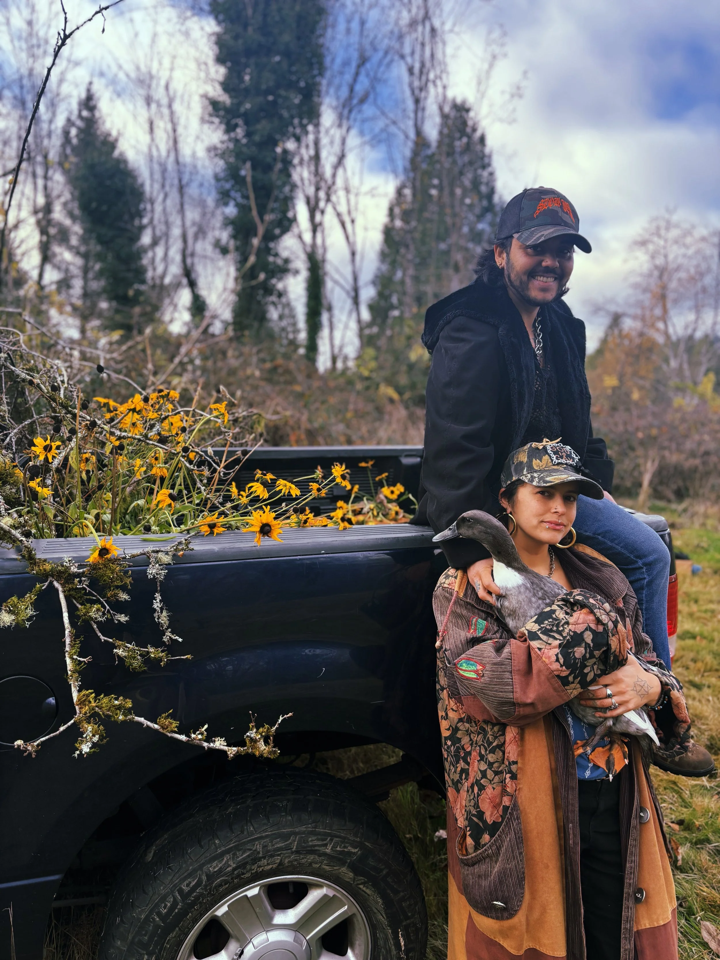 Two people outdoors near a truck filled with yellow flowers. One person is sitting on the truck bed, and the other is standing beside it, holding a duck. The background features trees and an overcast sky.