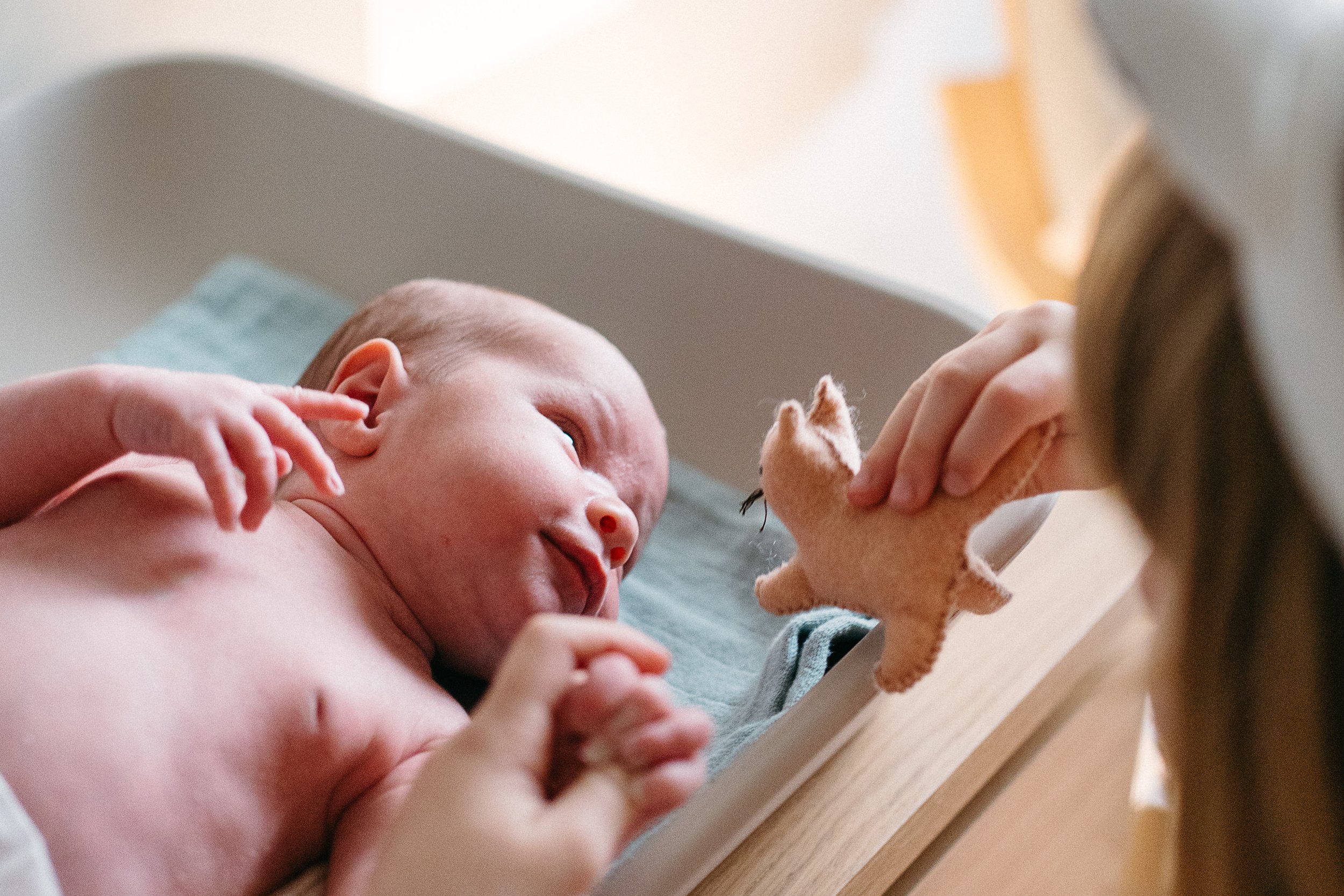 Baby lying on a changing table with a toy in front of him, being gently touched by a person.