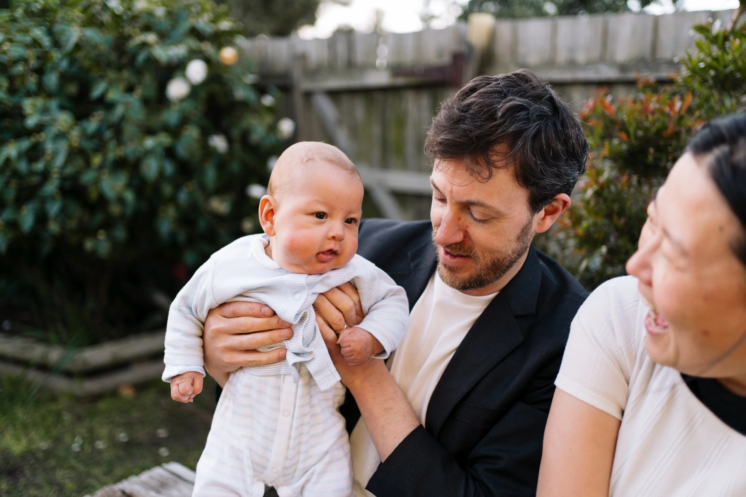 A man holding a baby outside next to a woman, all smiling and enjoying an outdoor moment near a wooden fence and greenery.