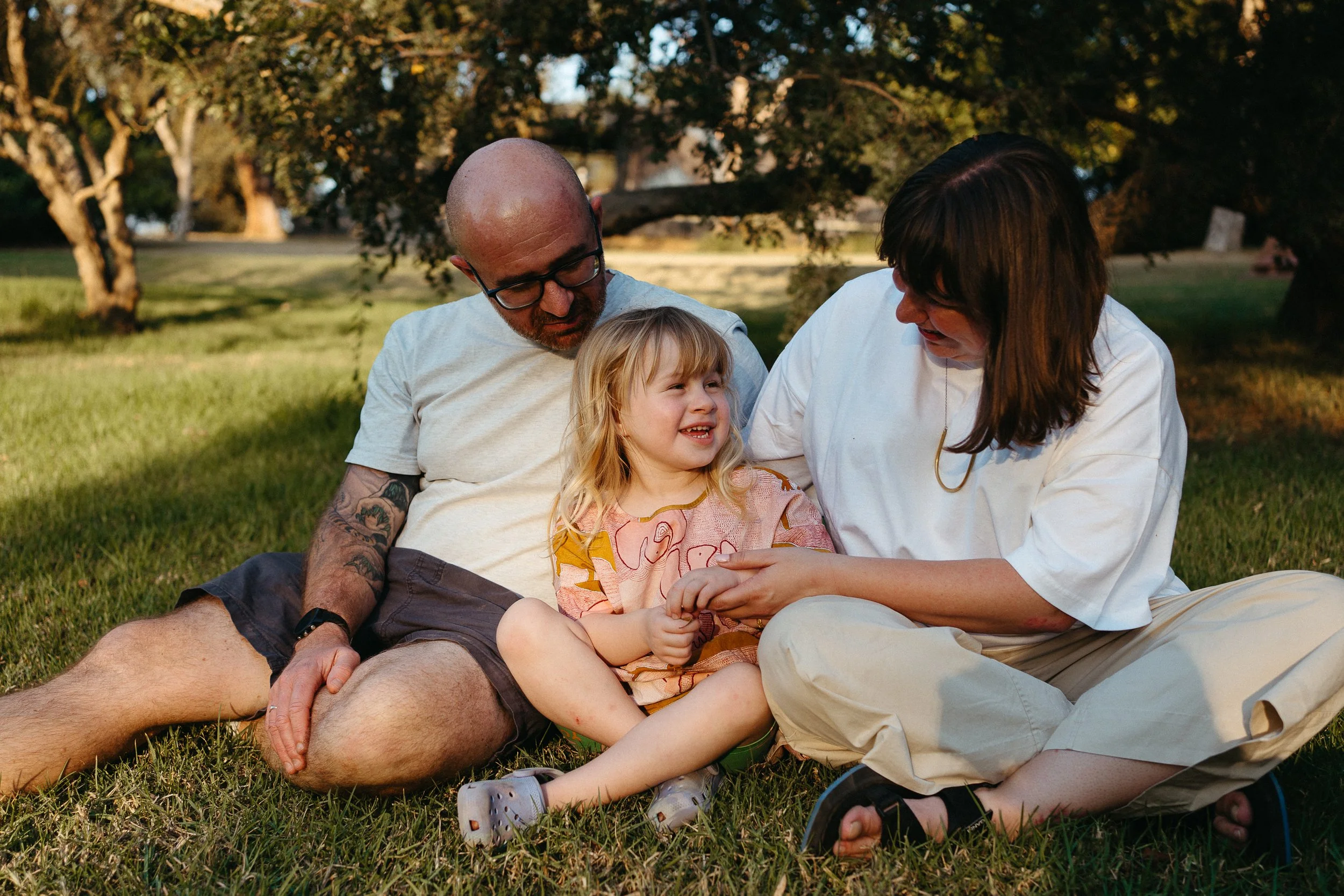 A family sitting on the grass in a park, a man, woman, and a young girl smiling and holding hands, the man has glasses and tattoos, trees in the background.
