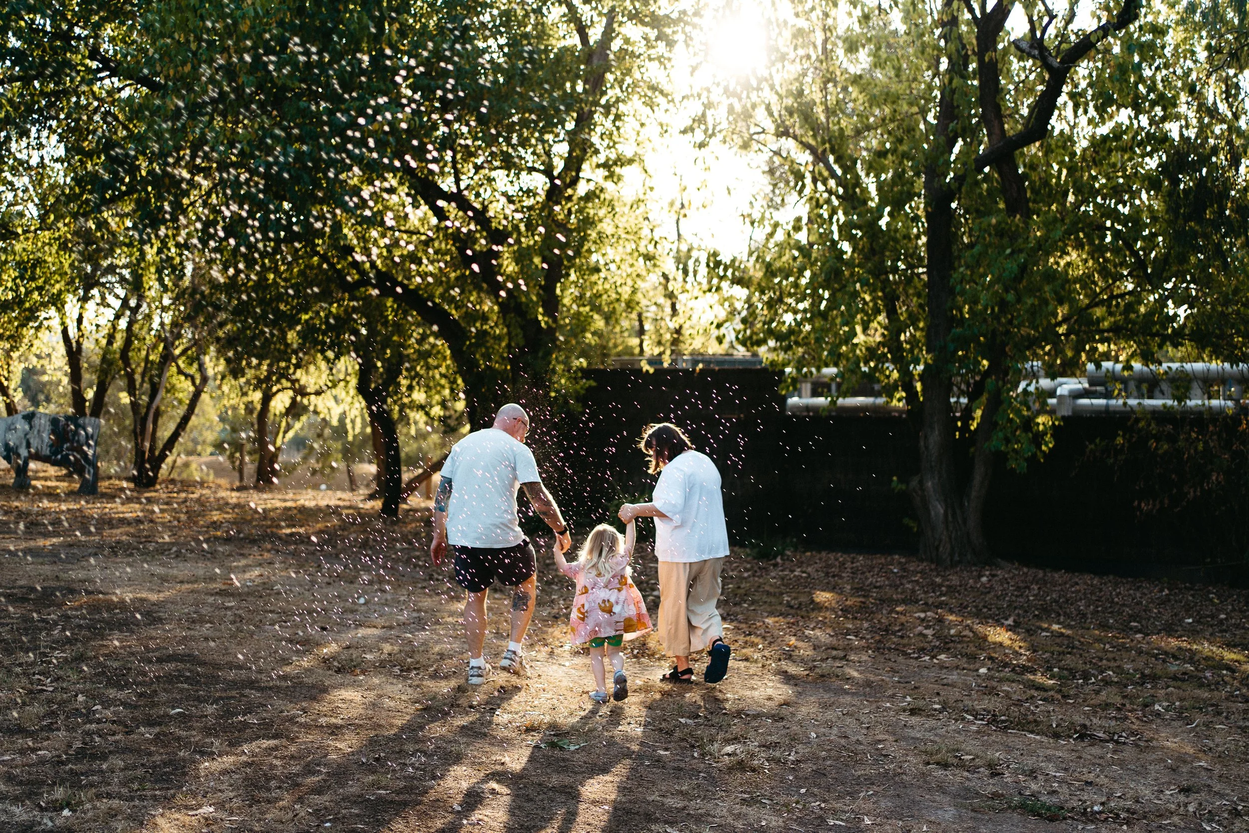 A family of three, including a man, woman, and young girl, playing with water spray in a park during late afternoon, with trees and sunlight in the background.