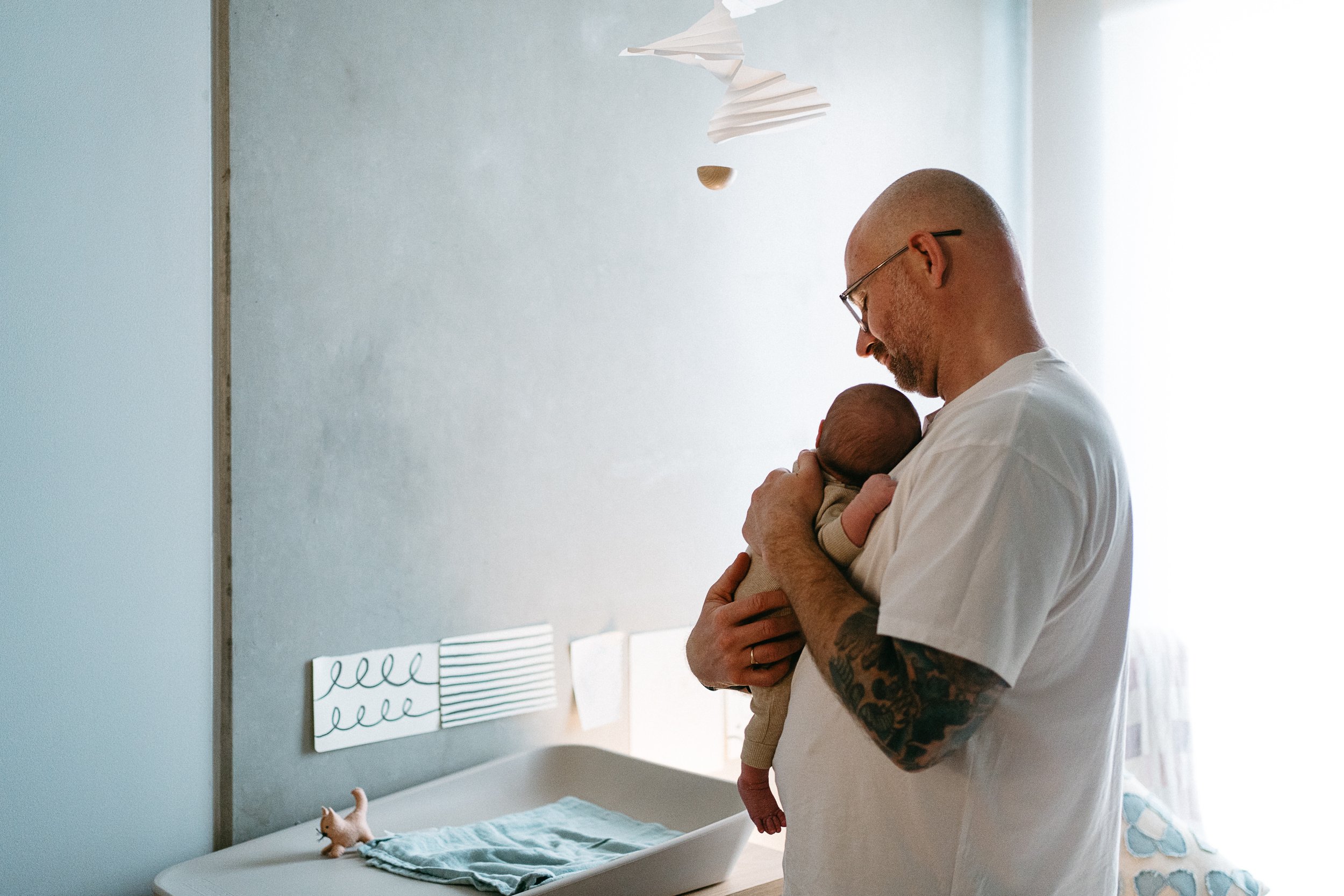 A man holding a newborn baby in a room with minimal decor, bright natural light, and a changing table with a plush toy pig on it.