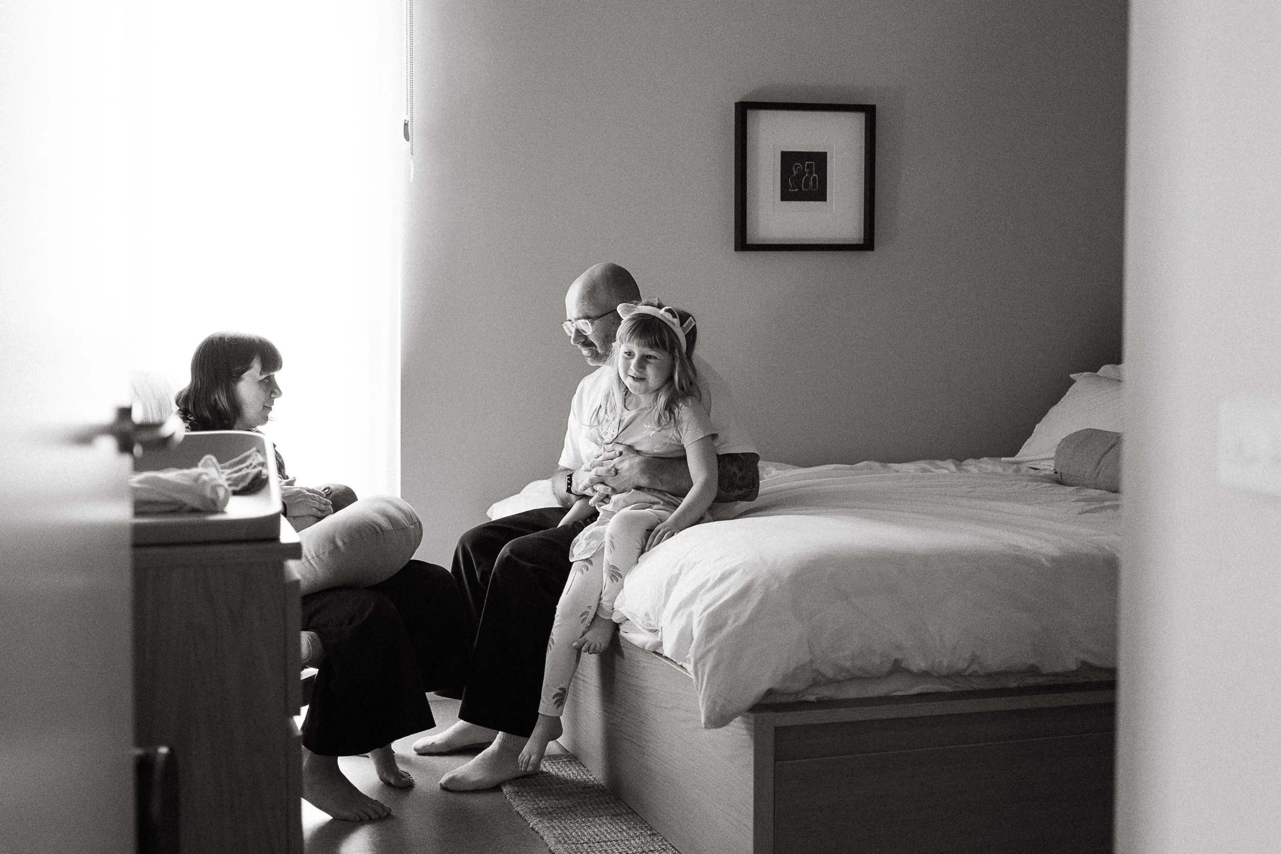 A man, a young girl, and a woman in a bedroom. The man is sitting on the bed with the girl on his lap, smiling. The woman is sitting on the floor facing them, engaging in a conversation. The scene is lit by natural light coming through a window.