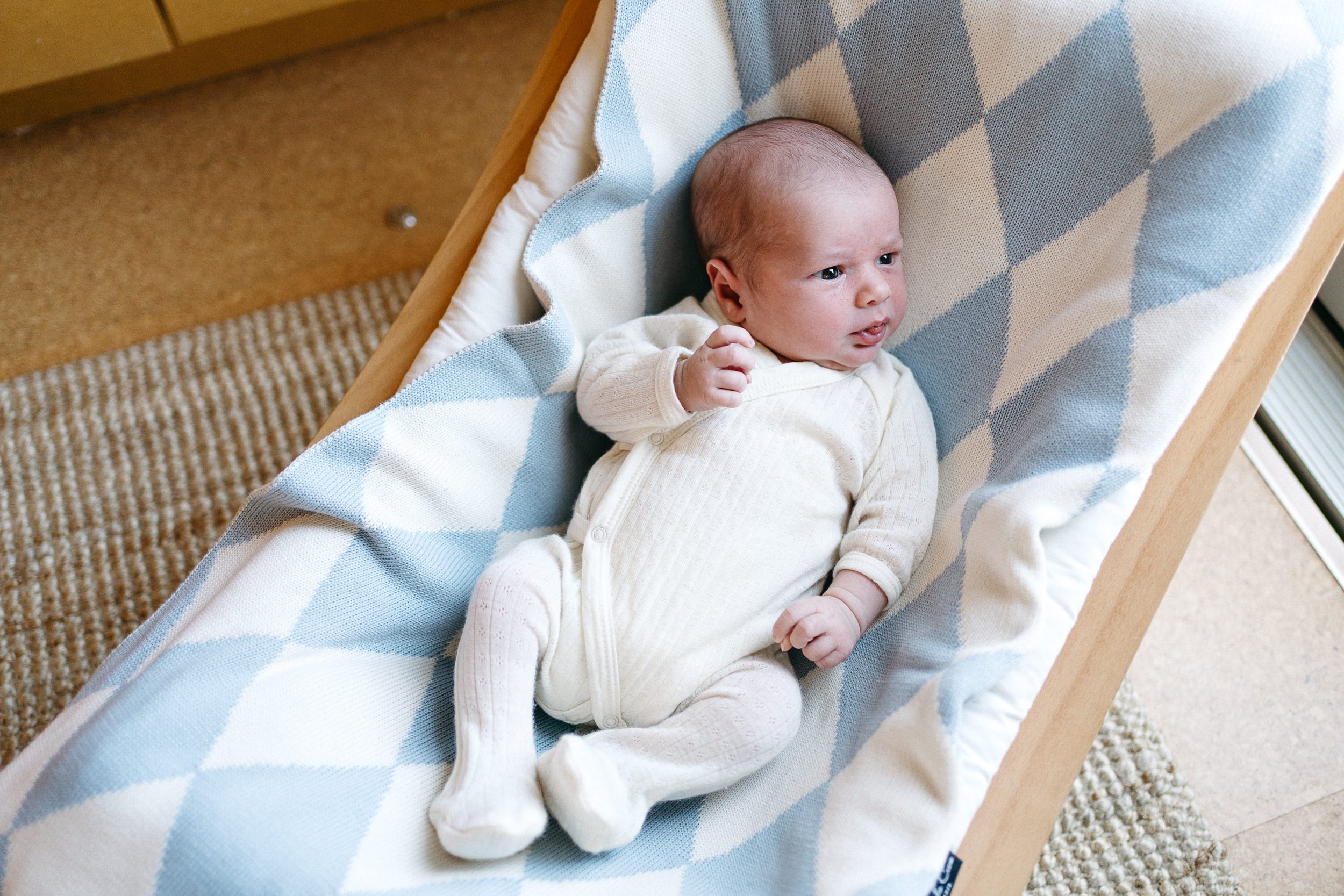 A baby lying in a wooden cradle with a blue and white checkered blanket, looking attentively to the side with a slightly furrowed brow.