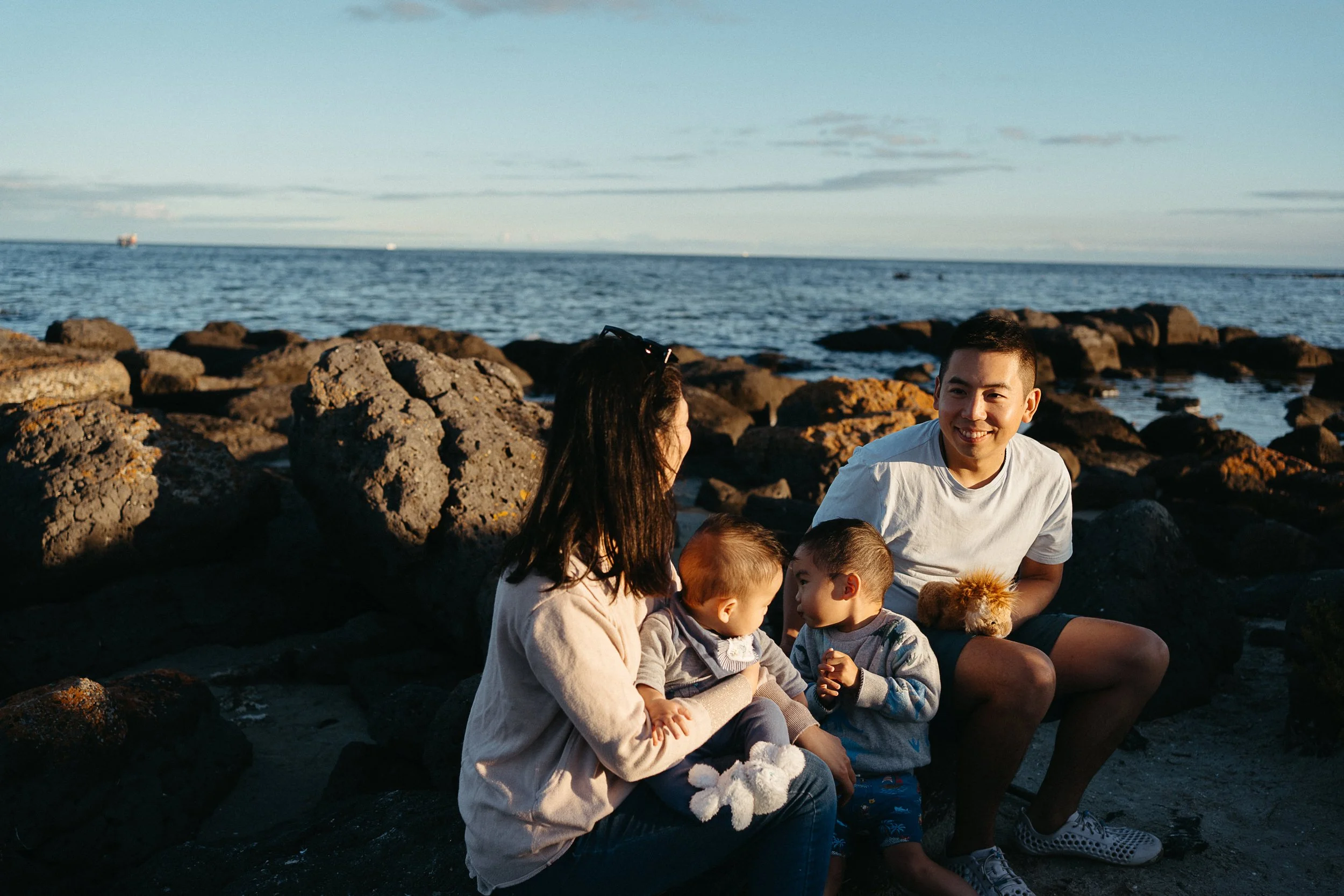 A family of four sitting on rocks by the ocean during sunset, smiling and engaging with each other.