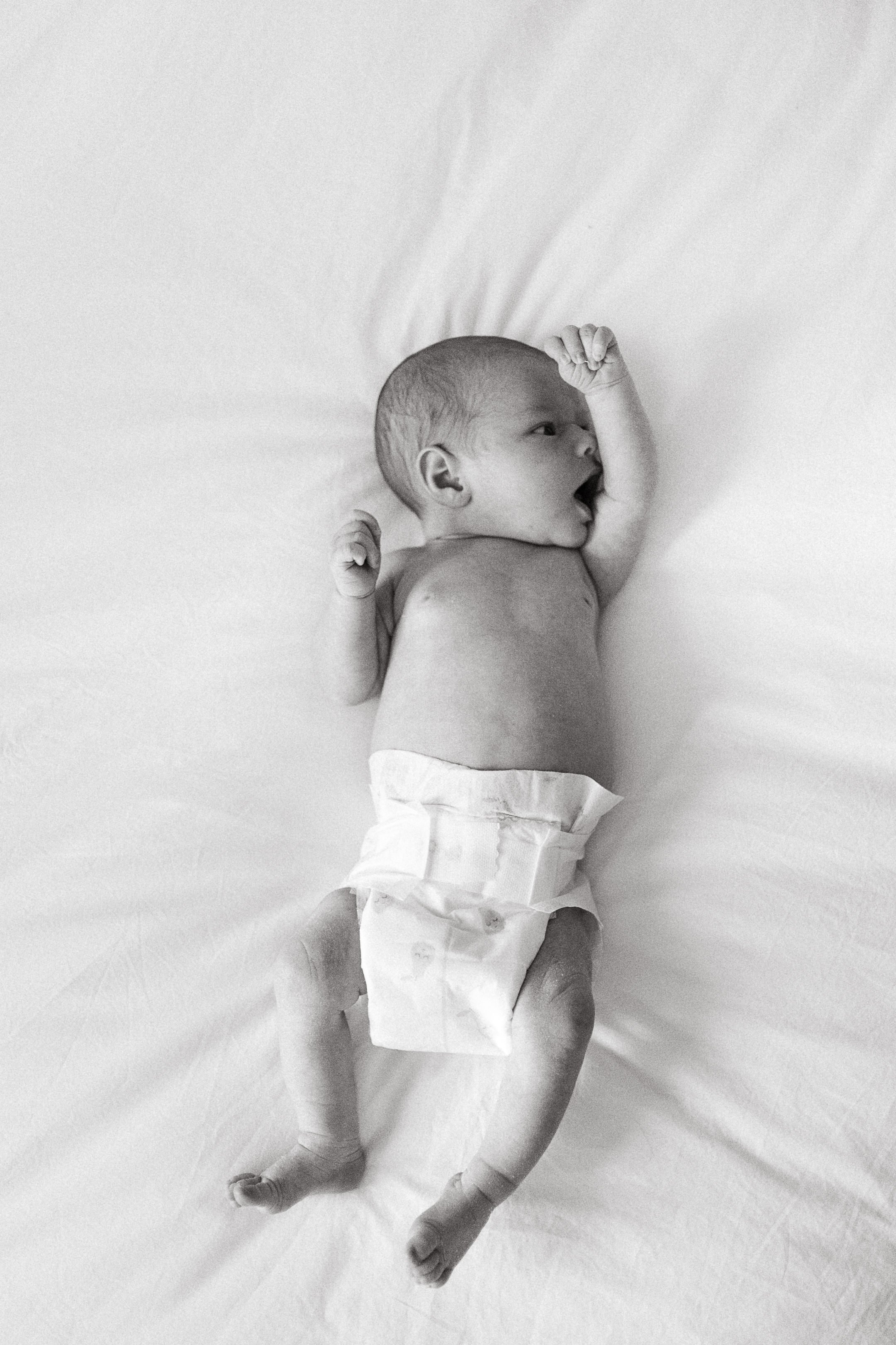 Black and white photo of a newborn baby lying on a white sheet, wearing a diaper, with one arm raised to the forehead and the mouth open.