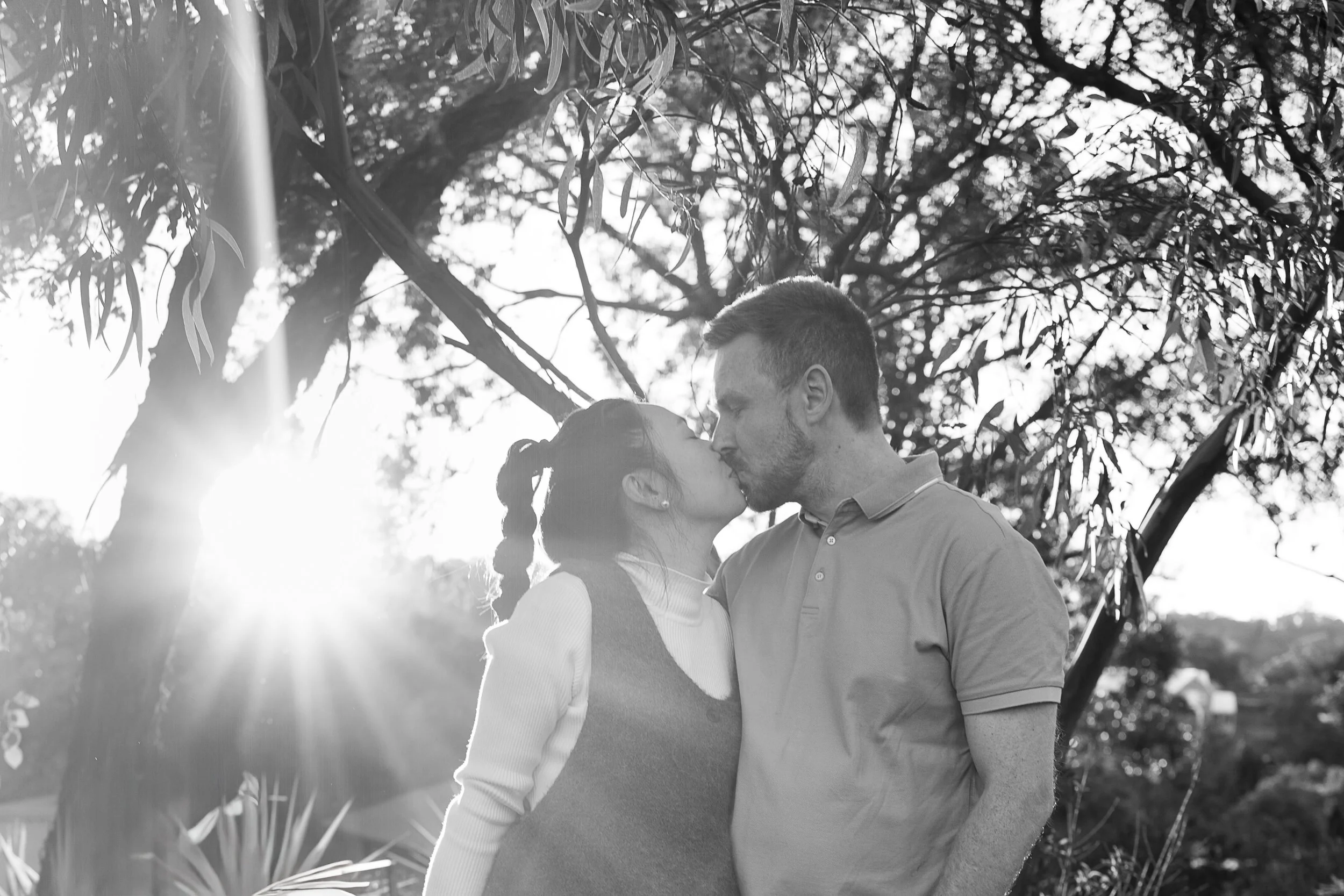 A couple kissing outdoors during sunset, with trees and sunlight in the background.