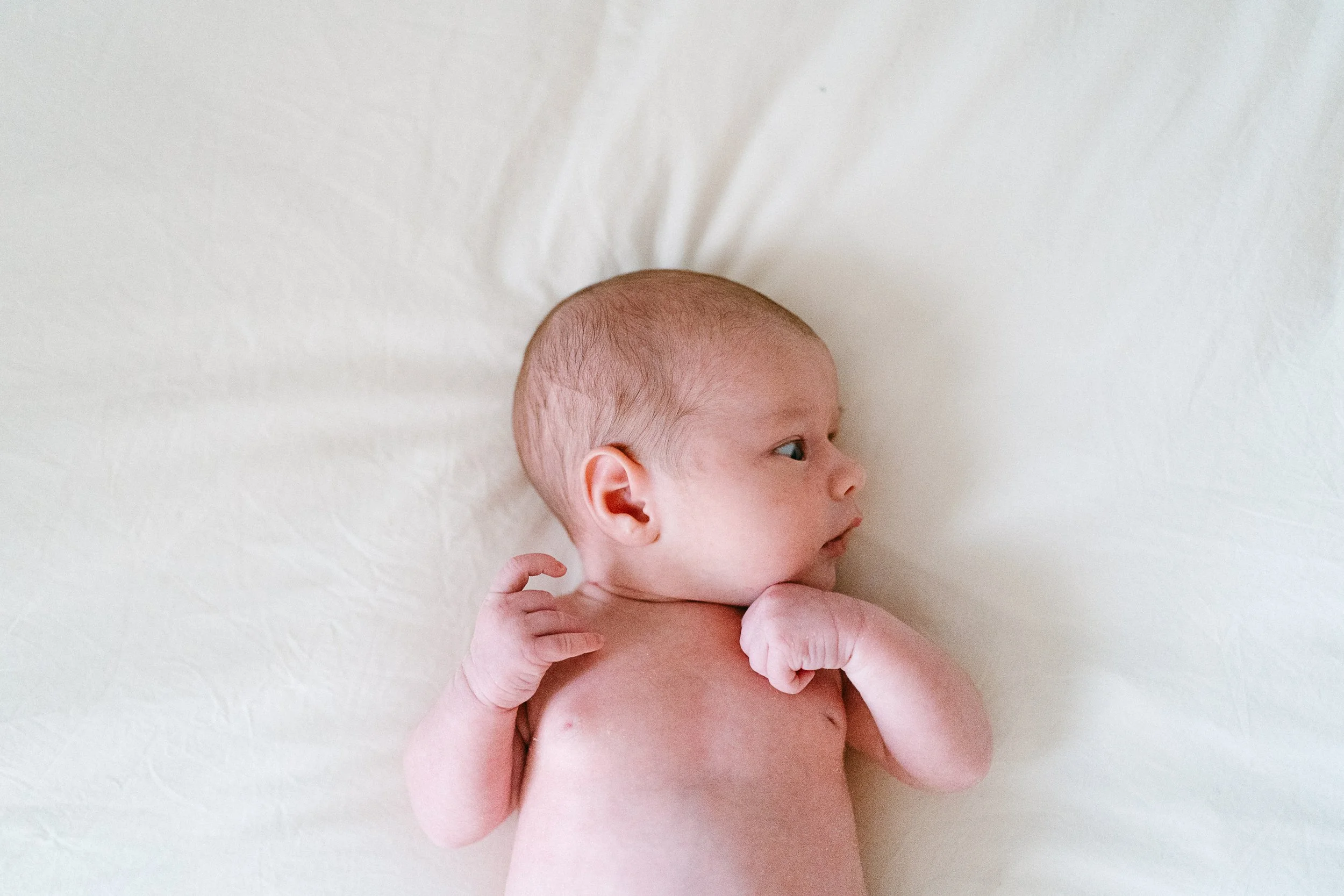 Newborn baby lying on a soft white surface, looking to the side with hands near face.