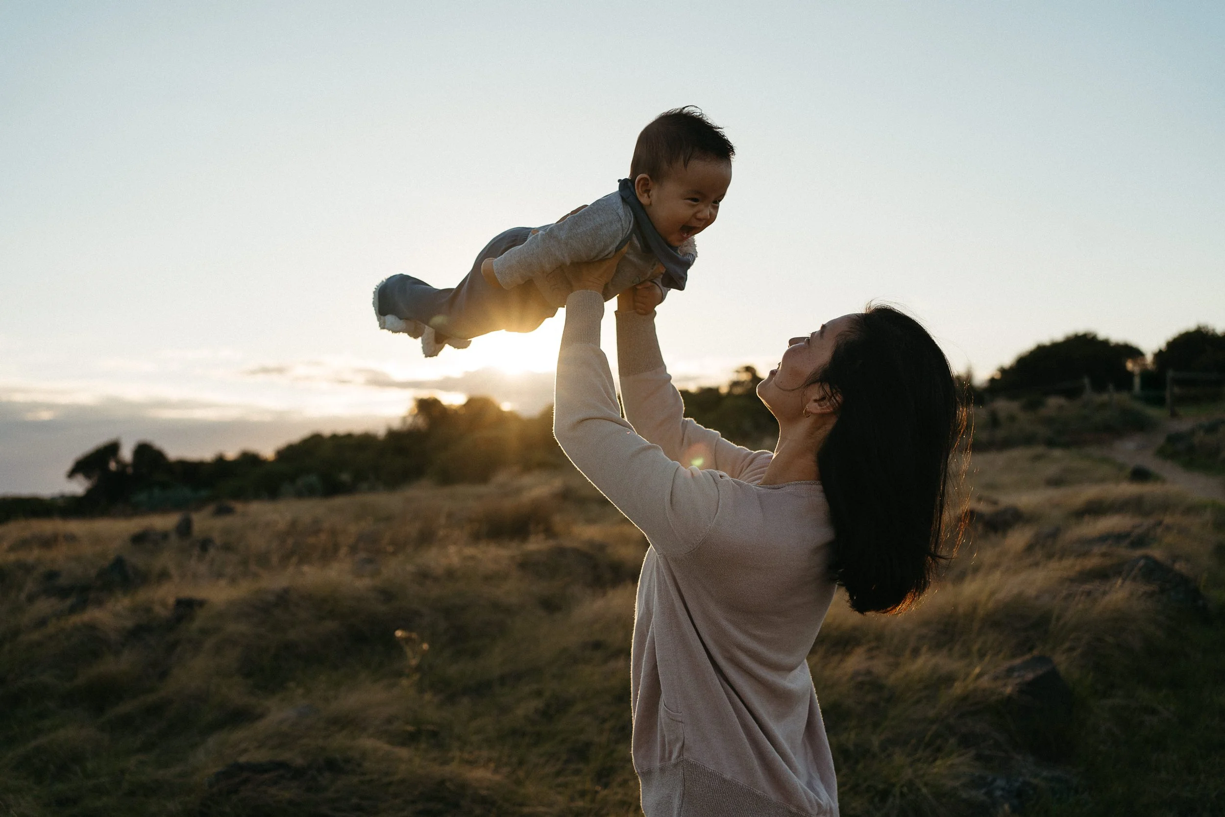 A woman lifting a young boy into the air outdoors at sunset, with a landscape of grass and trees in the background.