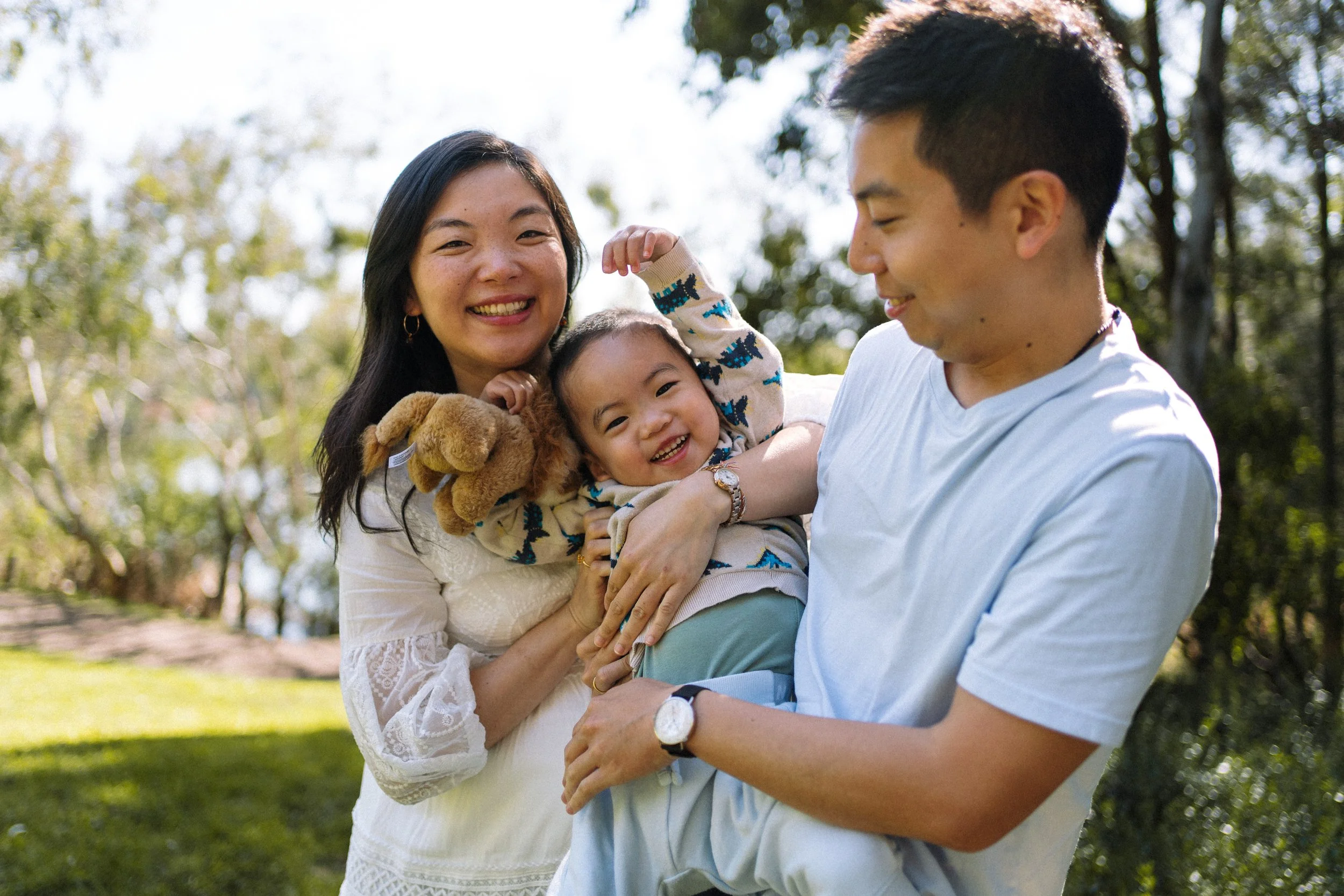 Family of three, including a woman, a man, and a young girl, smiling and playing together outdoors in a park on a sunny day.