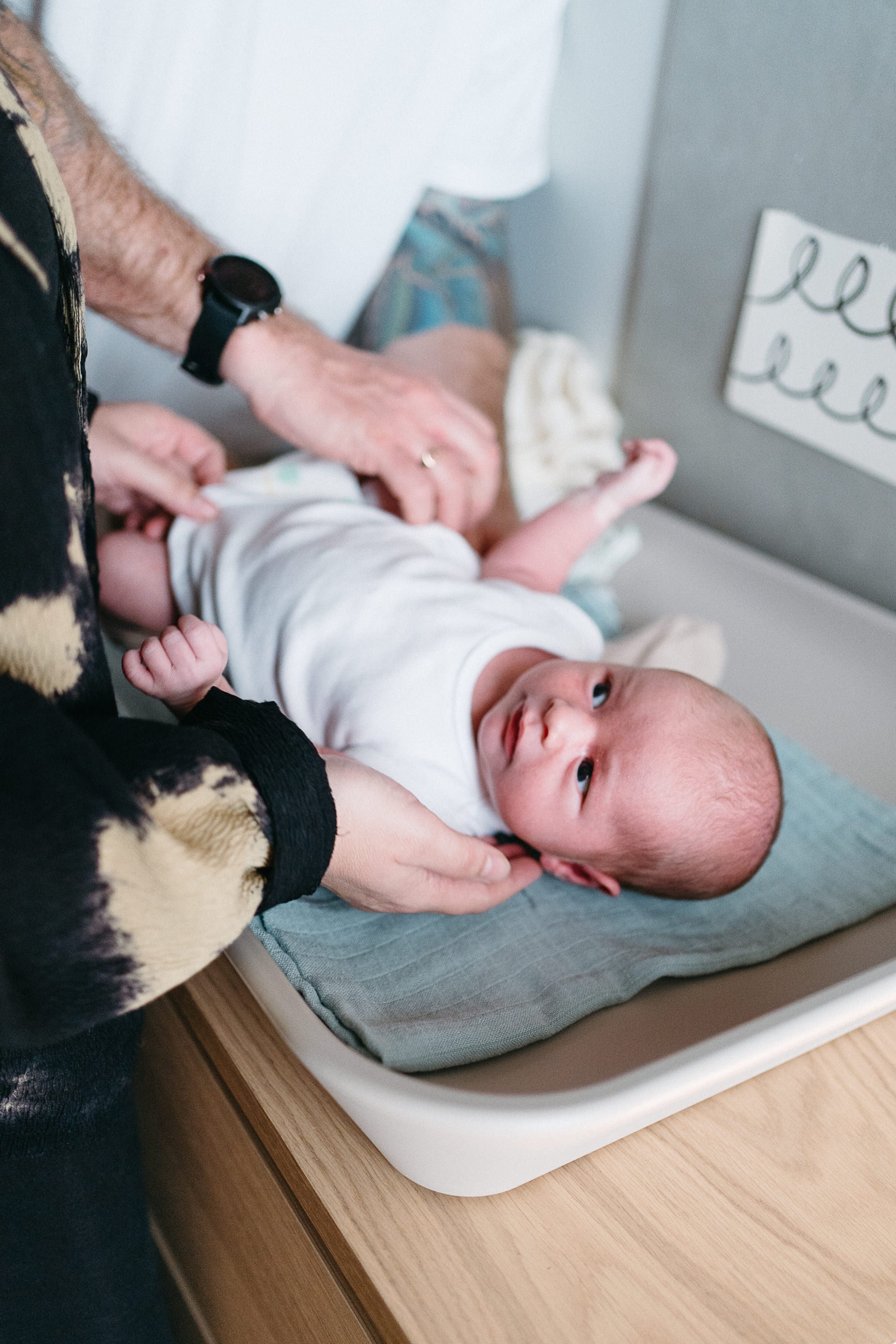 A newborn baby lying on a changing pad, looking up at the camera with a slight smile, being attended to by an adult.