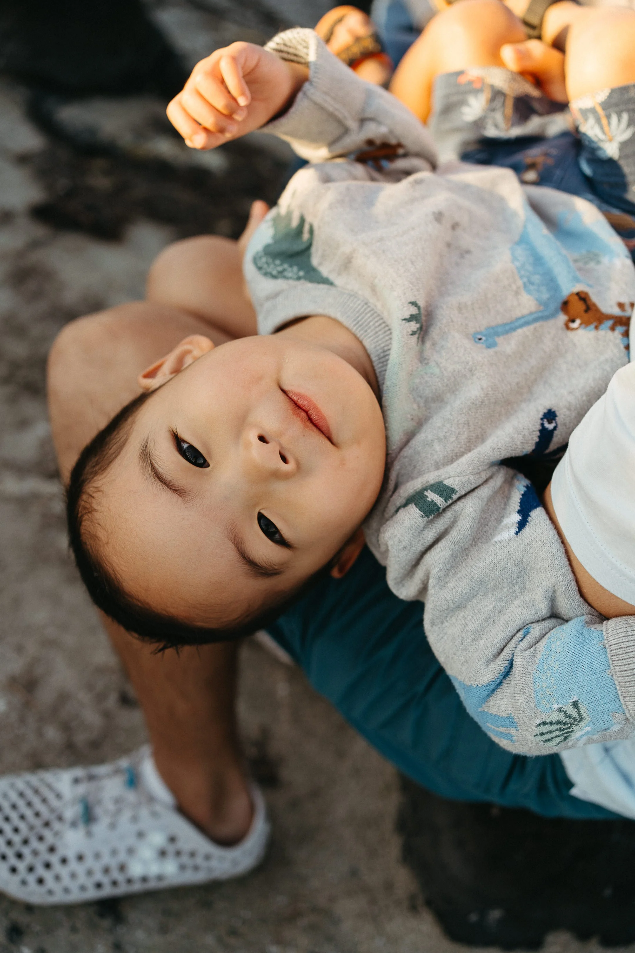 A young boy with short dark hair lying on a person's lap on a sandy surface, wearing a gray sweatshirt with animal patterns, looking up at the camera with a gentle expression.