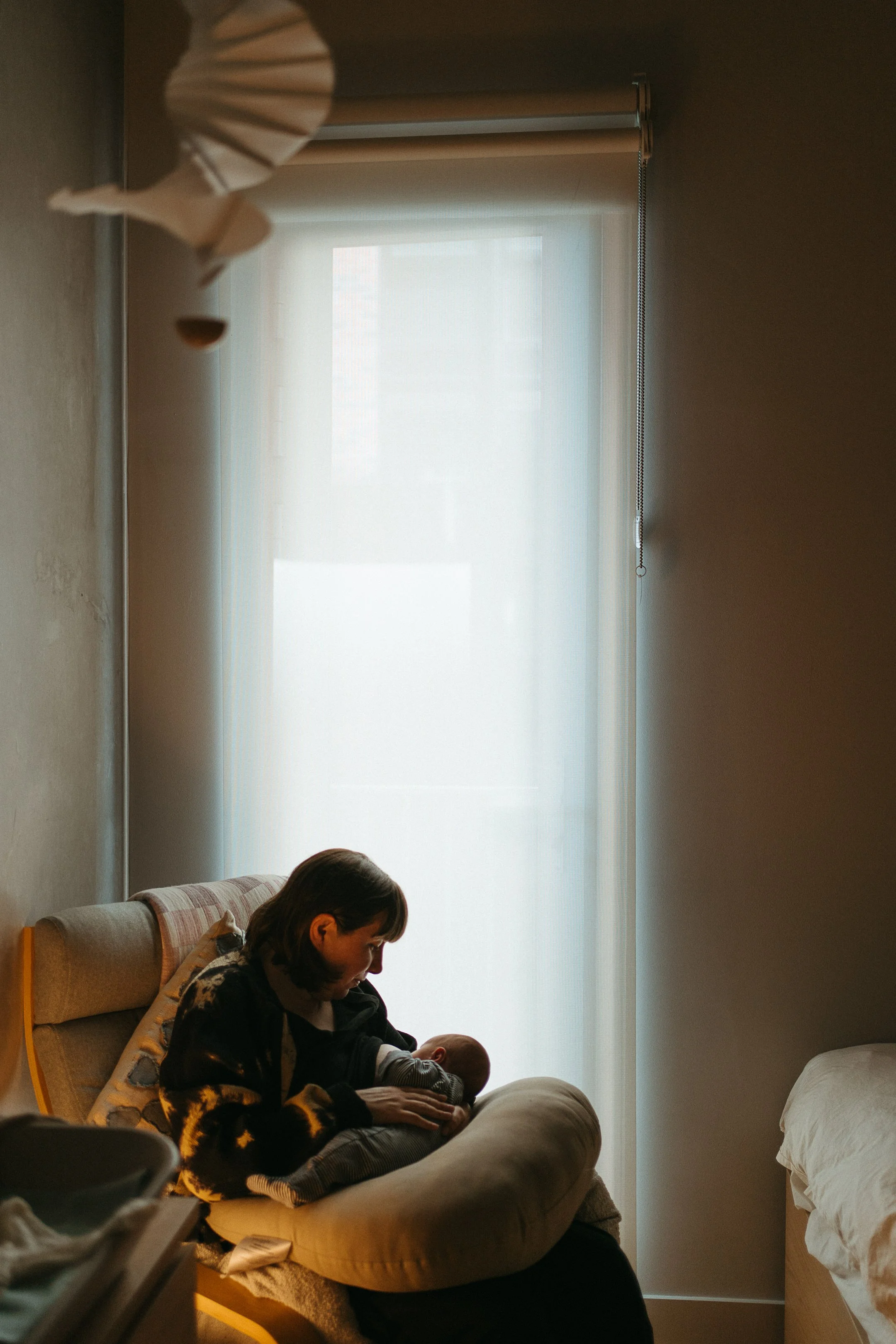 A woman sitting on a couch, holding and breastfeeding a baby near a window with sheer curtains.