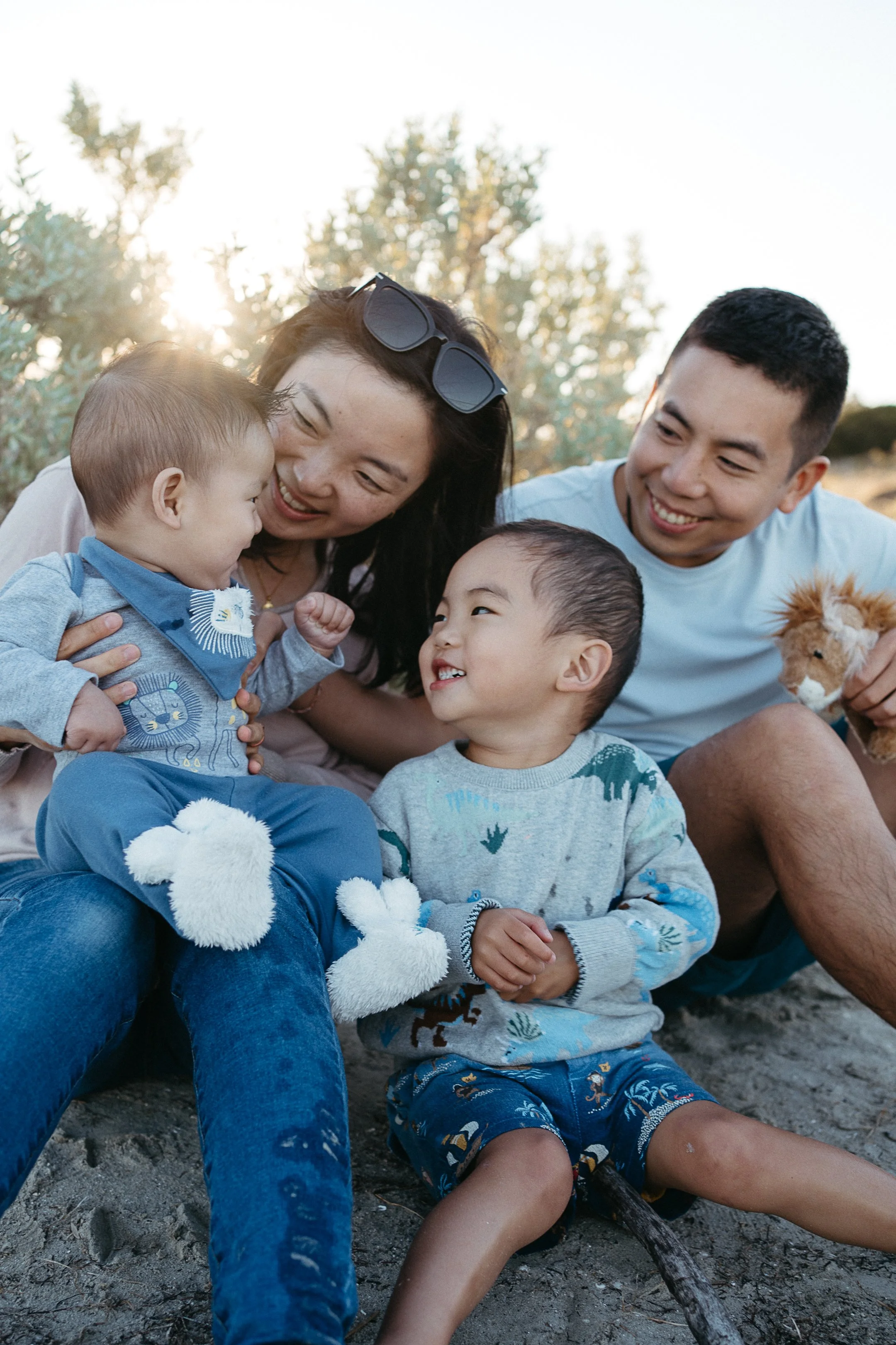 Family of four sitting on the ground outdoors, smiling and playing with two young kids, with trees and sunlight in the background.